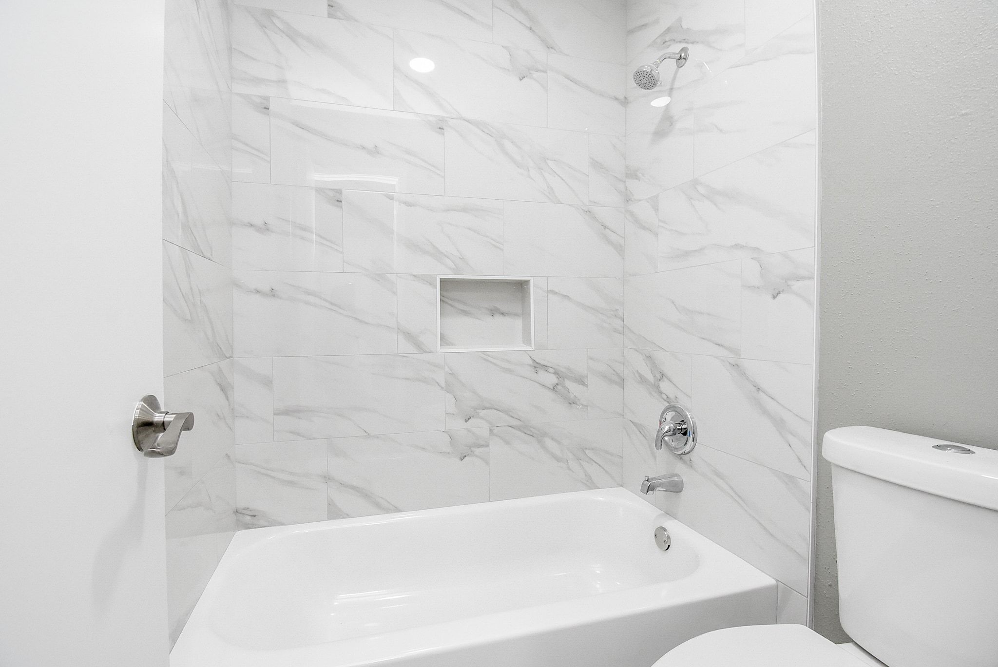 17330 Modbury Drive Spring, TX 77379 - Photo 18 of 30 Modern bathroom featuring elegant marble-style tiles, a sleek showerhead, and a pristine white bathtub with a convenient built-in shelf.