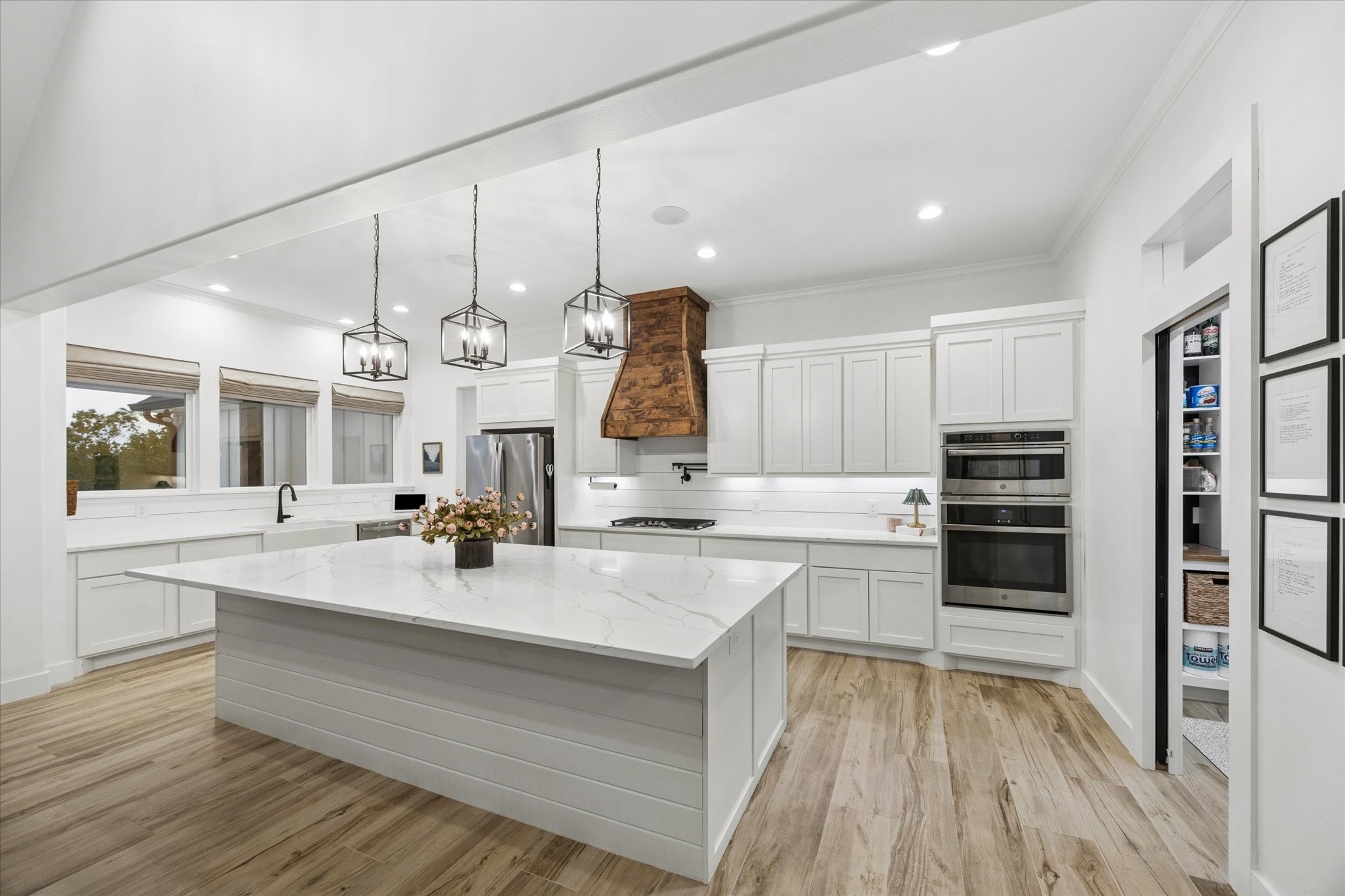 5131 Timber Ridge Drive Sealy, TX 77474 - Photo 13 of 50 Kitchen features a spacious layout with a large marble island, modern pendant lighting, and stainless steel appliances. White cabinetry, a wooden range hood, and light wood flooring add to its bright and contemporary feel, while a large pantry offers additional storage space.