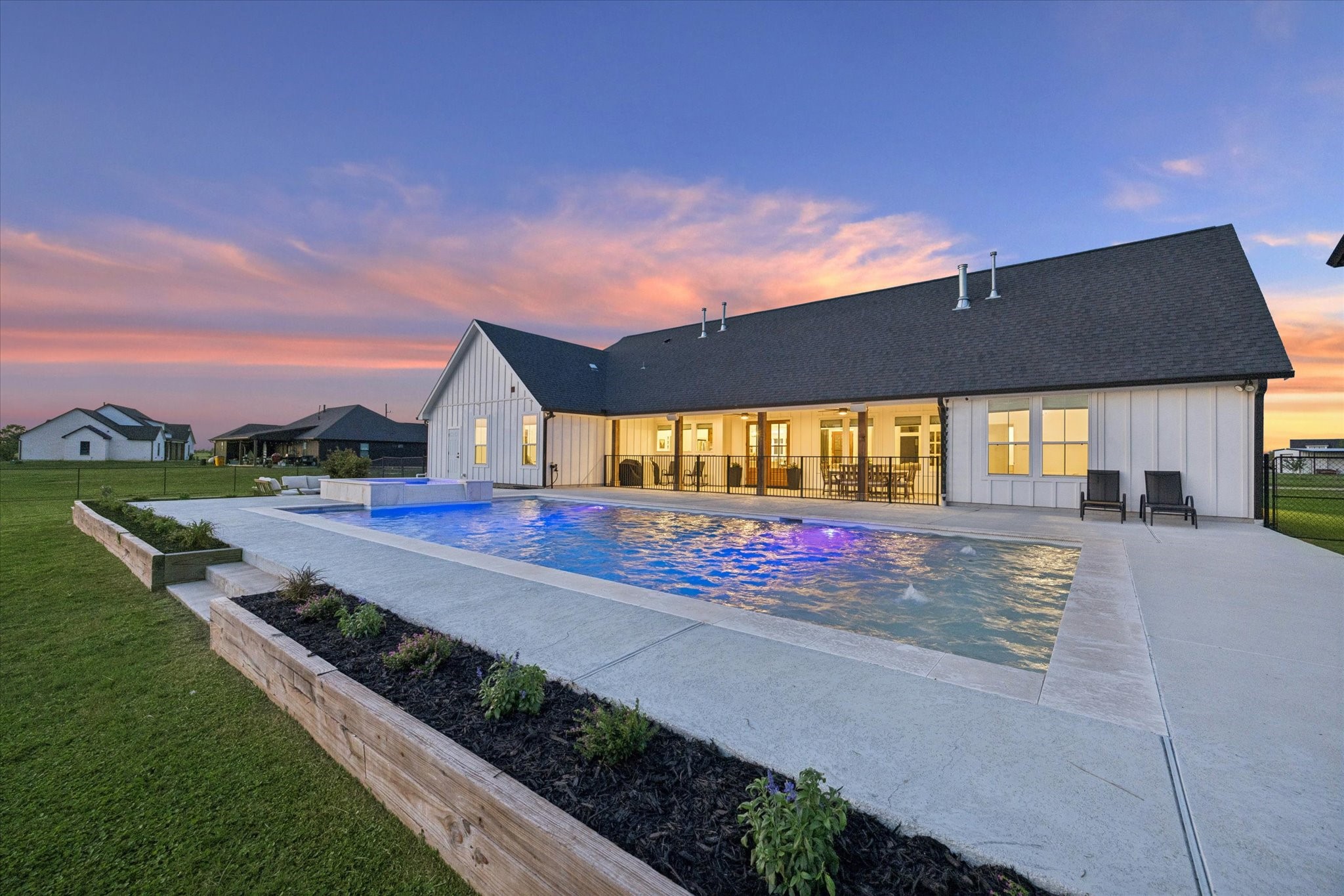 5131 Timber Ridge Drive Sealy, TX 77474 - Photo 45 of 50 This photo showcases a modern farmhouse-style home with a spacious backyard featuring a large swimming pool. The house has a covered patio, perfect for outdoor dining and relaxation. The sunset provides a beautiful backdrop, enhancing the serene and inviting atmosphere.