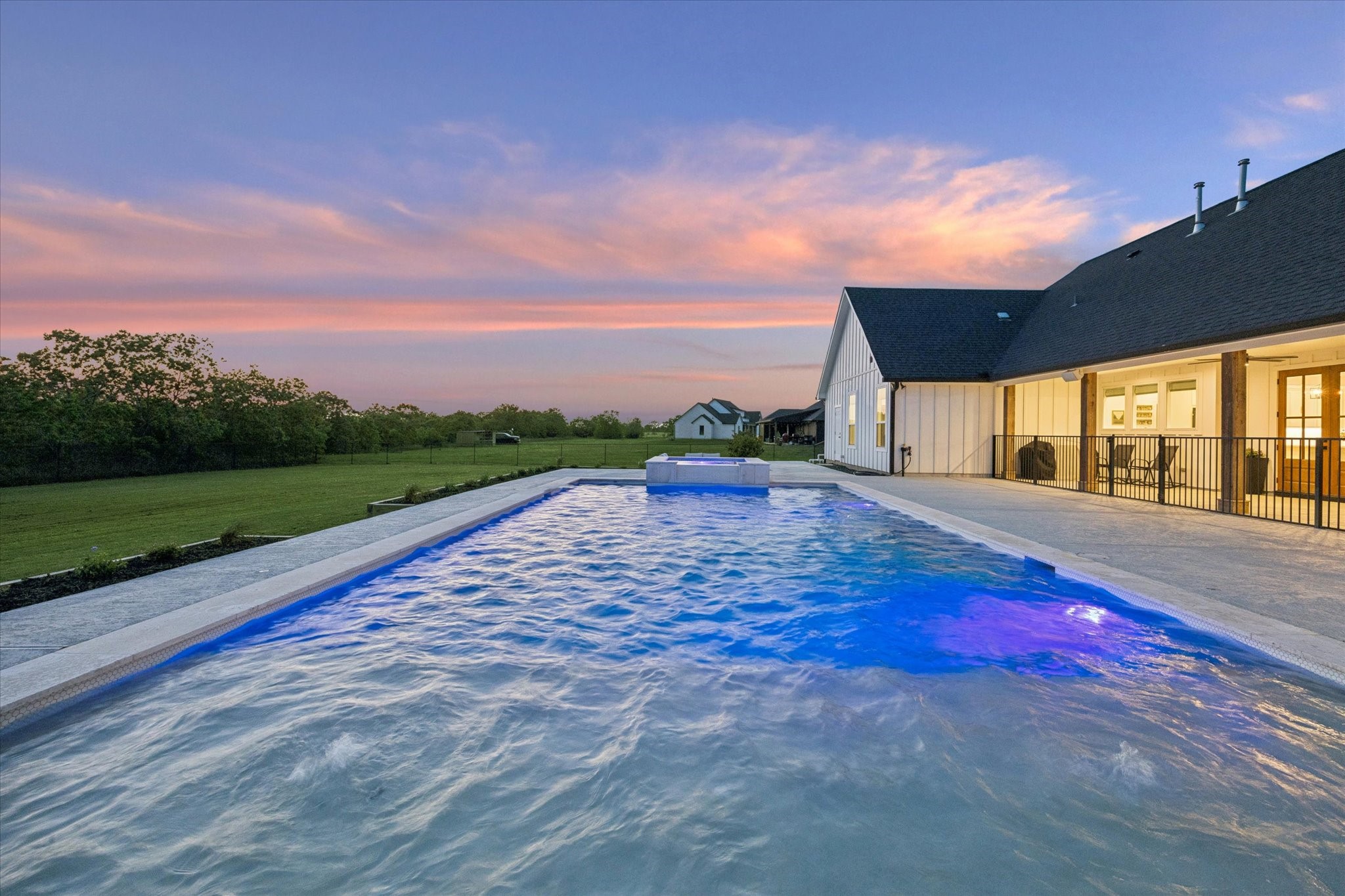 5131 Timber Ridge Drive Sealy, TX 77474 - Photo 46 of 50 This photo showcases a stunning backyard with a large, illuminated pool adjacent to a modern house. The expansive lawn and serene sunset create a welcoming, tranquil atmosphere.