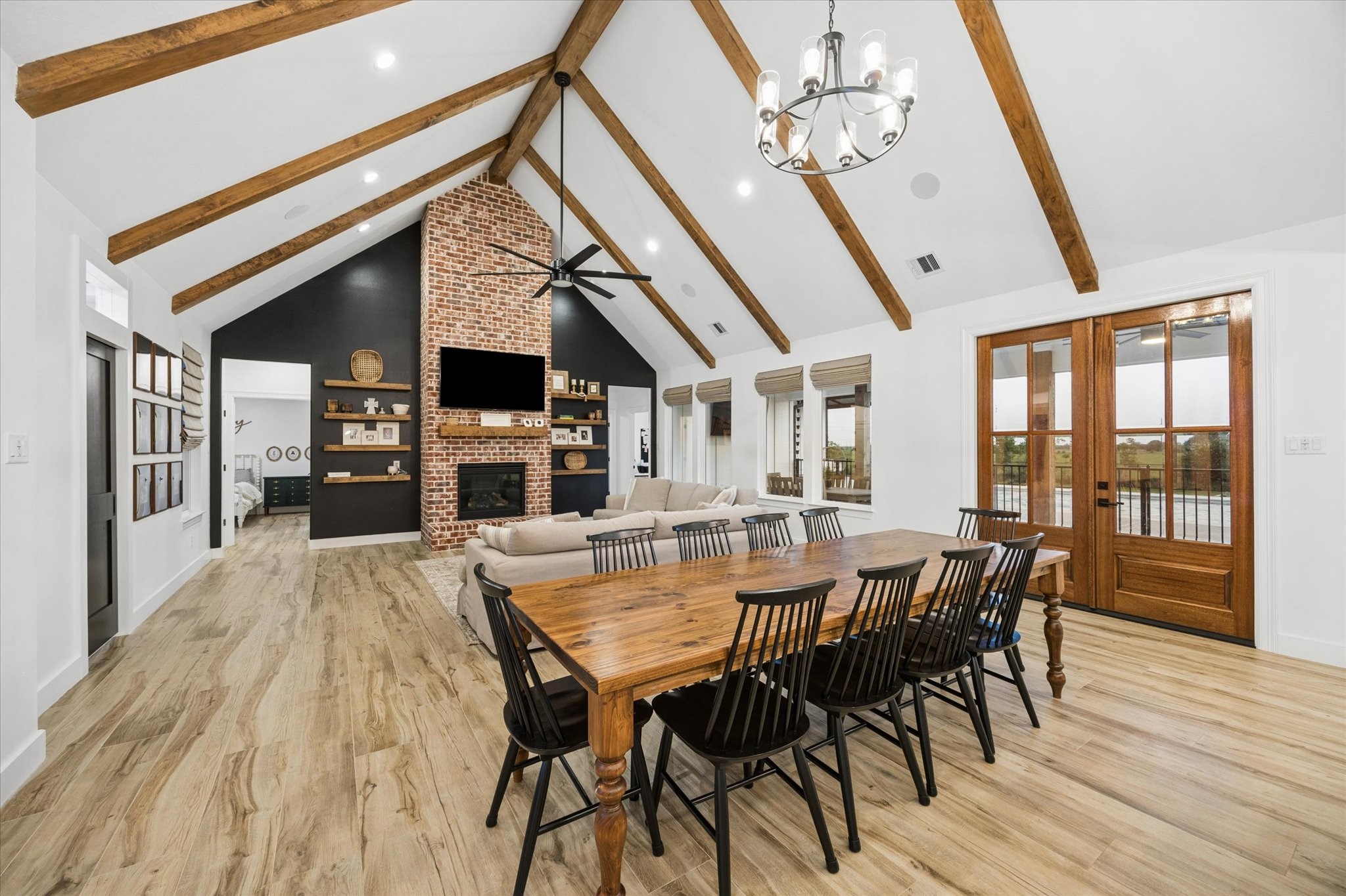 5131 Timber Ridge Drive Sealy, TX 77474 - Photo 6 of 50 This open-concept living space features high vaulted ceilings with exposed wooden beams, a striking brick fireplace, and a cozy seating area. The room is bright and airy, with large windows and double doors leading outside. A spacious dining area with a modern chandelier completes the inviting atmosphere.