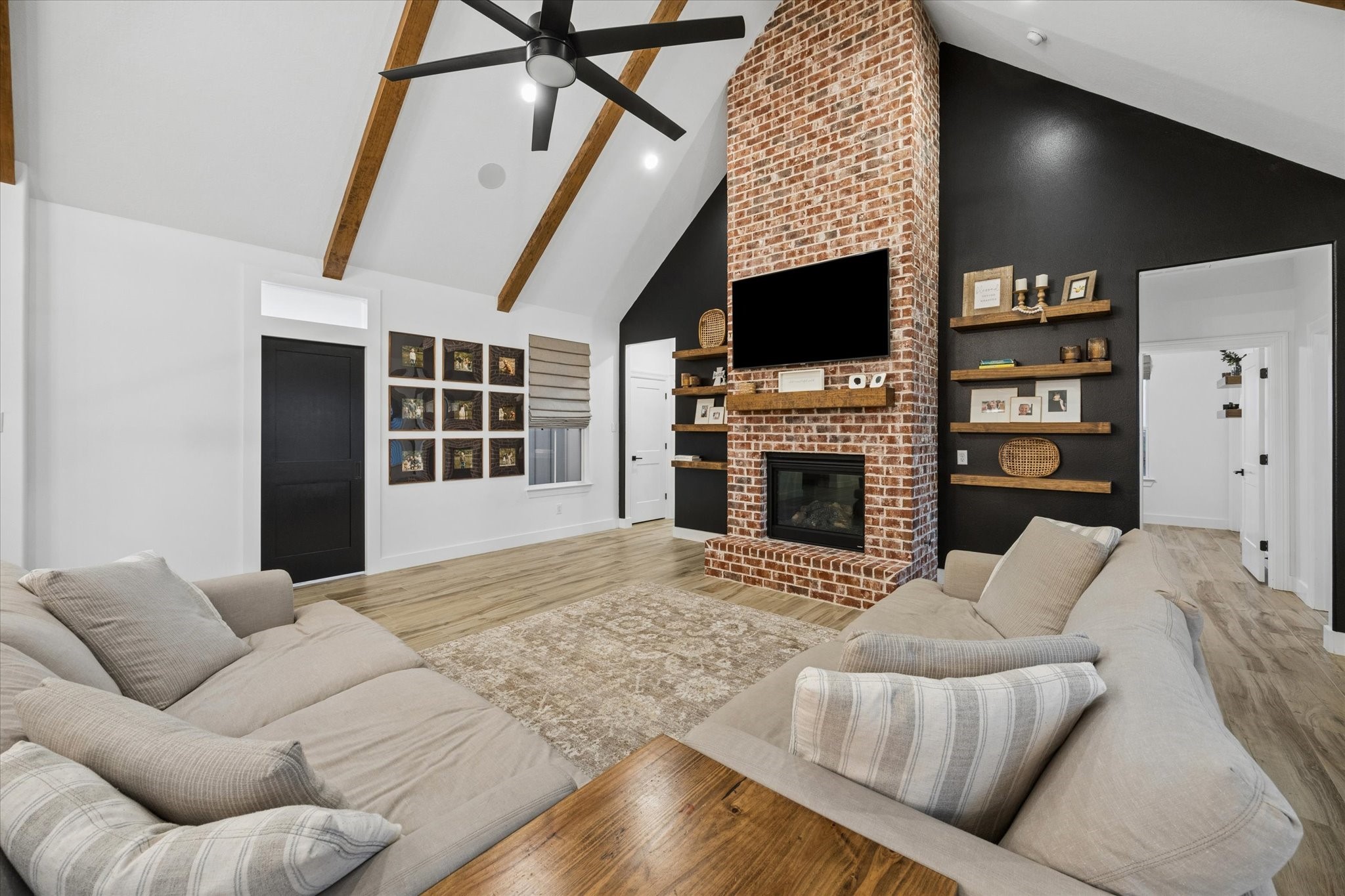 5131 Timber Ridge Drive Sealy, TX 77474 - Photo 9 of 50 This inviting living room features a high vaulted ceiling with exposed wooden beams and a striking floor-to-ceiling brick fireplace. The space is cozy with stylish built-in shelves, perfect for displaying decor. A large ceiling fan enhances comfort, while the open layout creates a welcoming atmosphere.
