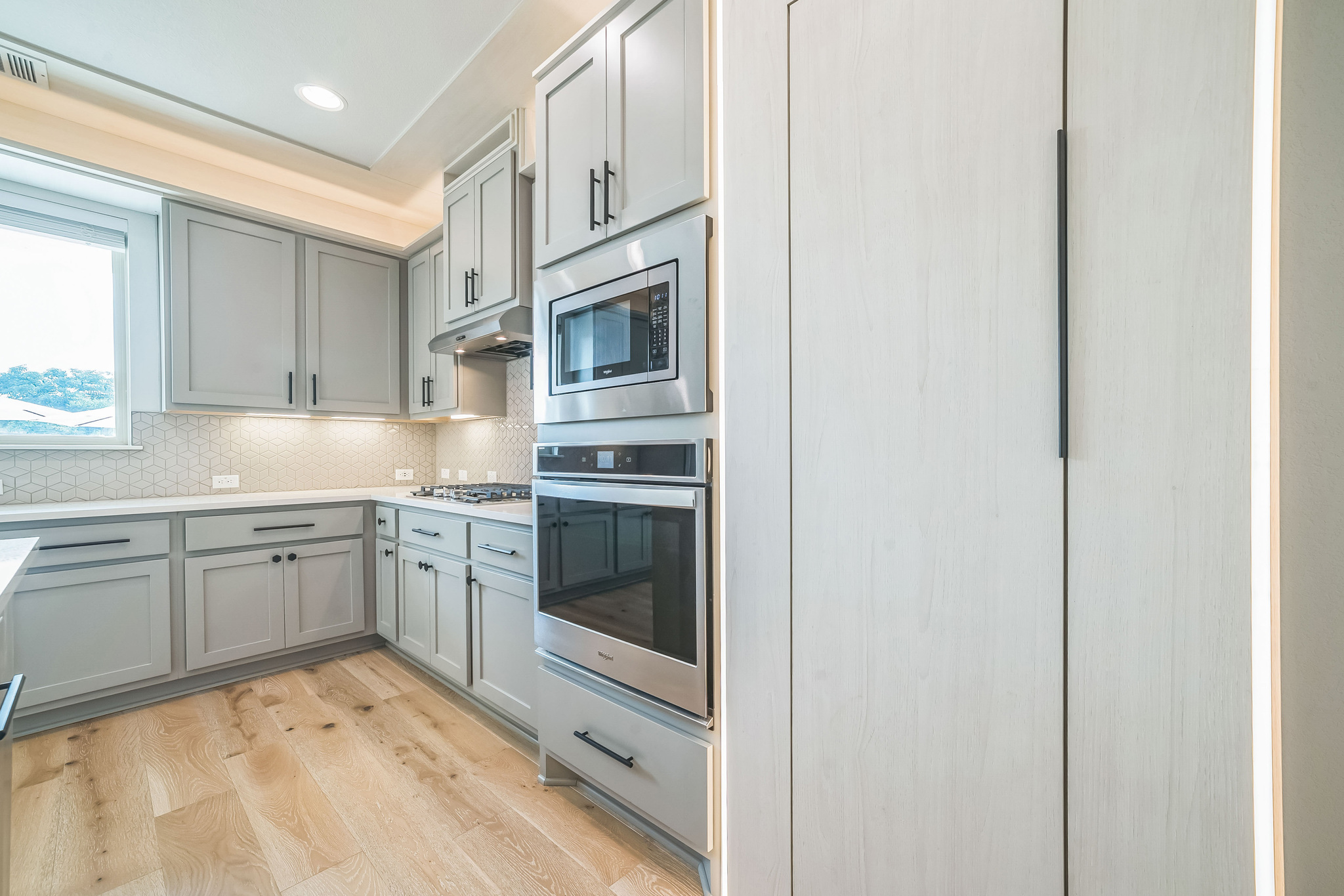14300 McCoy Loop, Unit 21 Austin, TX 78717 - Photo 11 of 40 The kitchen features light wood flooring, gray cabinetry with dark hardware, a white geometric tile backsplash, and stainless steel appliances including a built-in oven and microwave