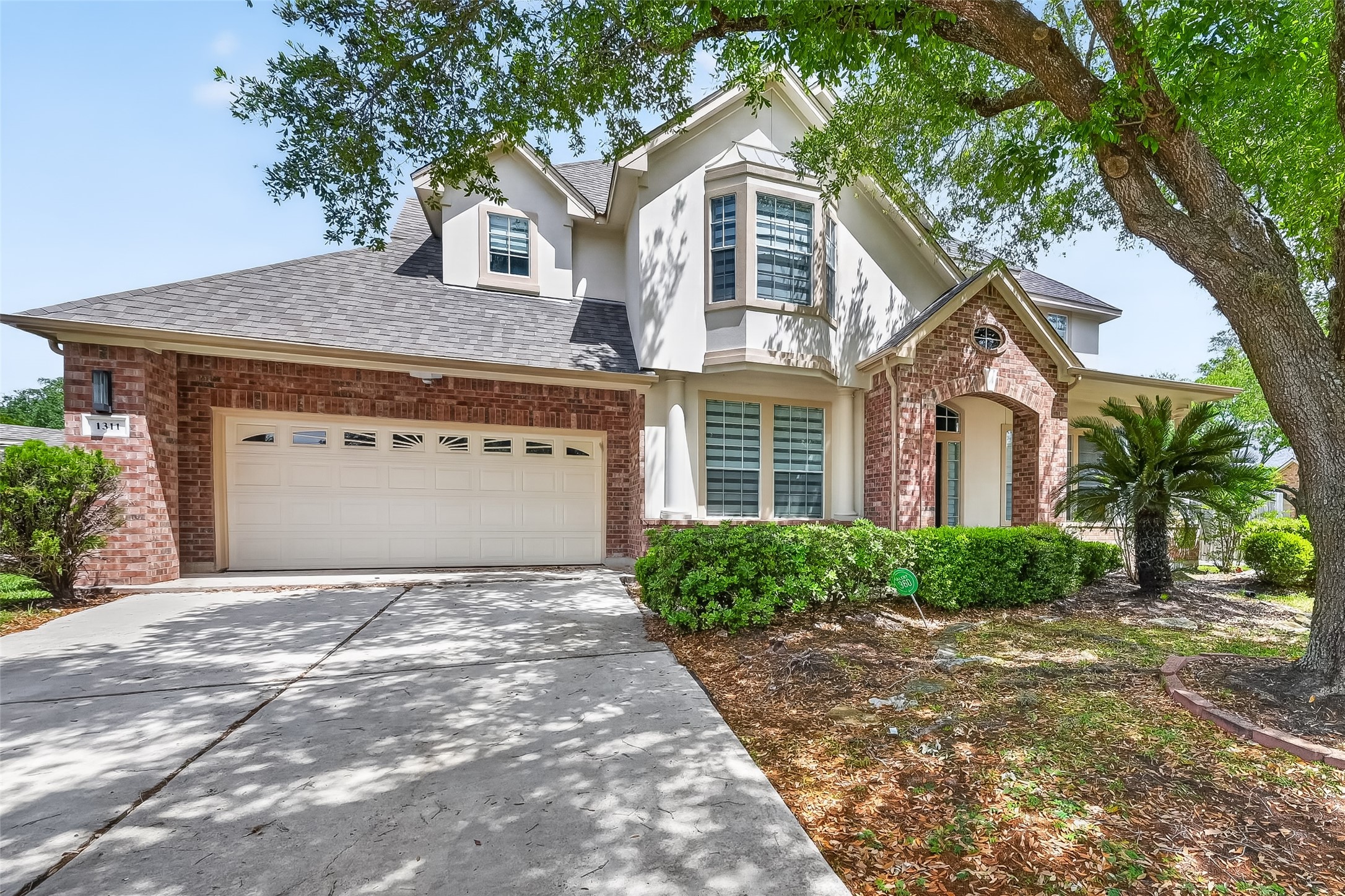 This photo shows a two-story house with a brick and stucco exterior. It features a two-car garage, large windows, and a welcoming front entrance. The property is surrounded by mature trees and landscaped greenery, offering a pleasant curb appeal.