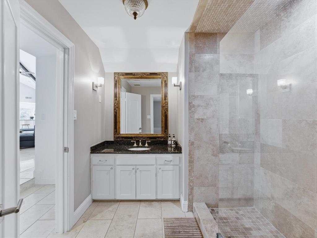6160 Spring Valley Road Dallas, TX 75240 - Photo 23 of 40 Spacious bathroom featuring a glass-enclosed shower with stone tile, a vanity with a dark countertop and gold-toned fixtures, and a decorative framed mirror