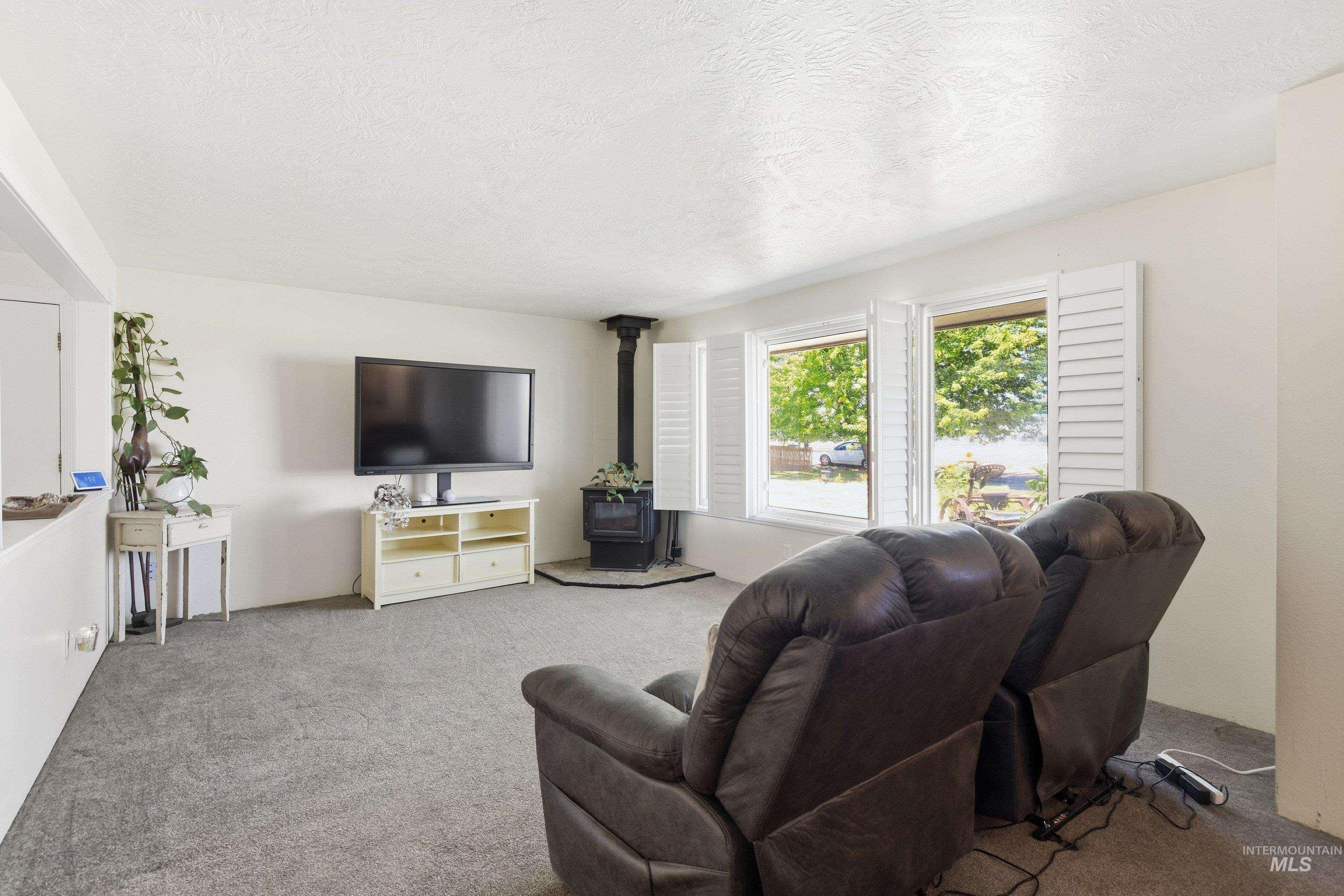 15095 David Street Caldwell, ID 83607 - Photo 9 of 39 Spacious living area featuring soft gray carpeting, a corner wood-burning stove with a hearth, and multiple windows with white plantation shutters