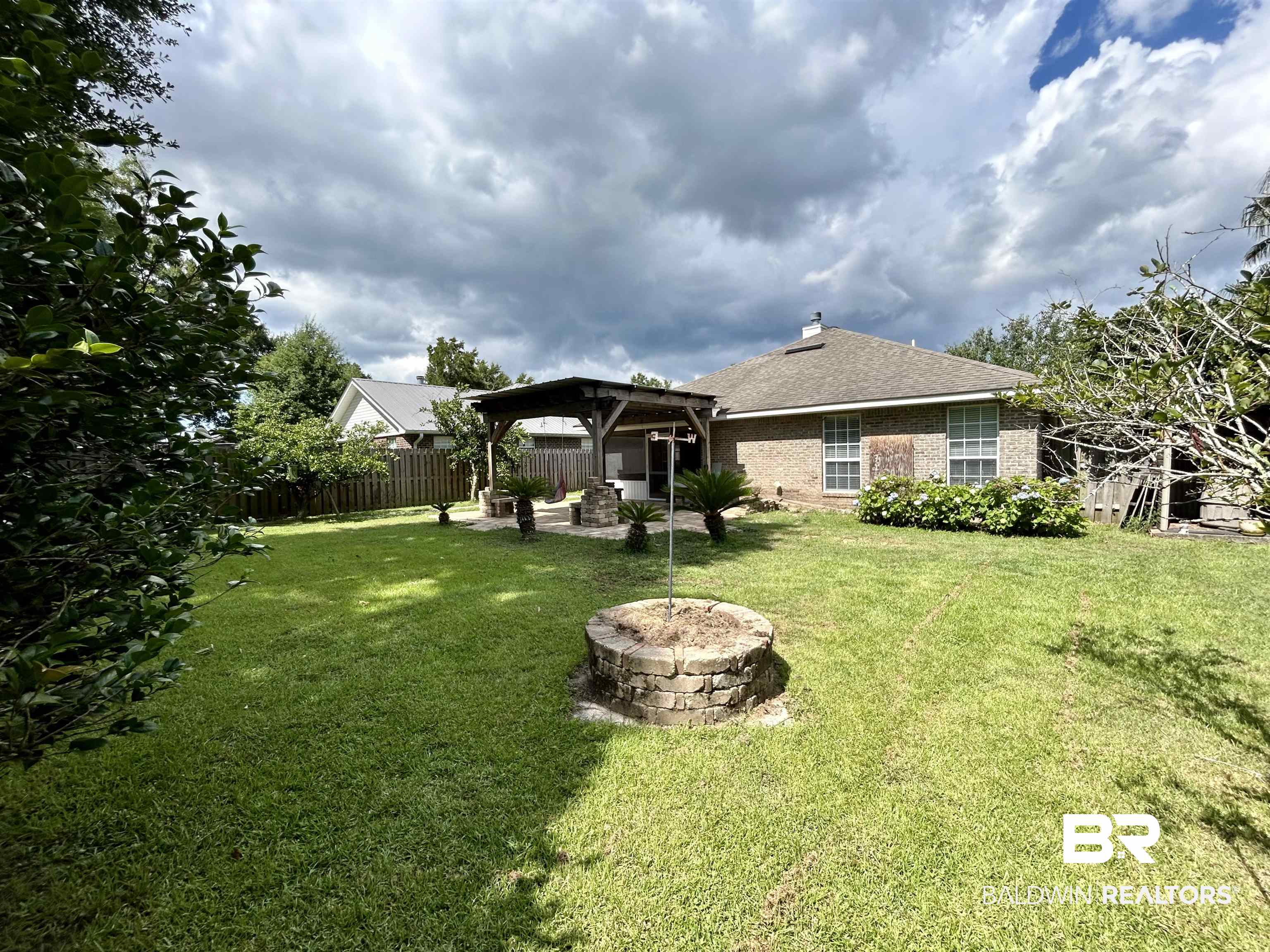 7198 Raintree Lane Gulf Shores, AL 36542 - Photo 33 of 33 Expansive backyard featuring a stone-clad fire pit, a covered patio with a wood frame, brick exterior, and mature perimeter foliage