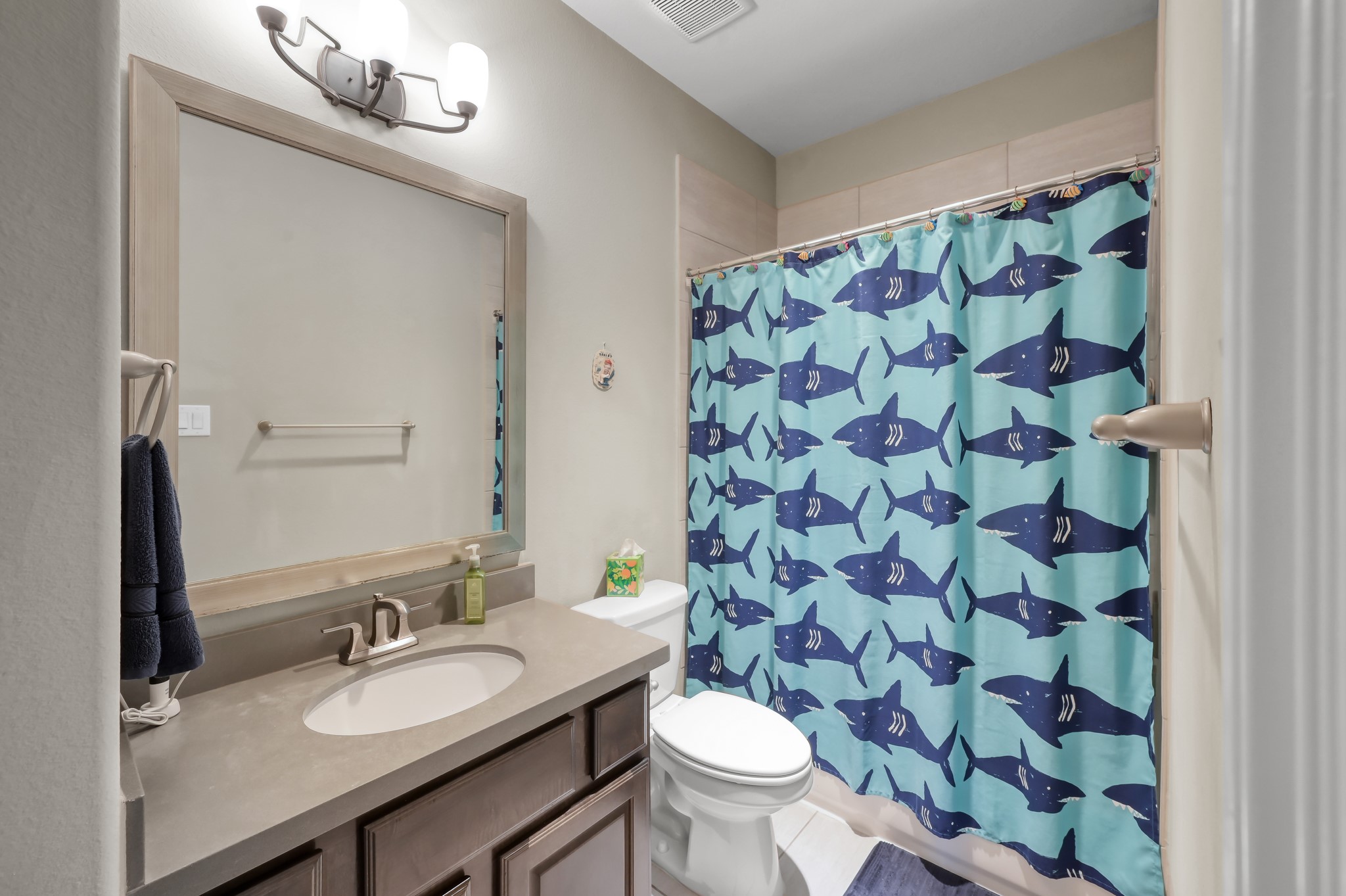 26 Woodborough Way Spring, TX 77389 - Photo 26 of 43 This bathroom in one of the bedrooms upstairs, features a modern vanity with a large mirror, stylish lighting, and a playful shark-themed shower curtain. It has a neutral color palette and is well-lit, creating a welcoming atmosphere.