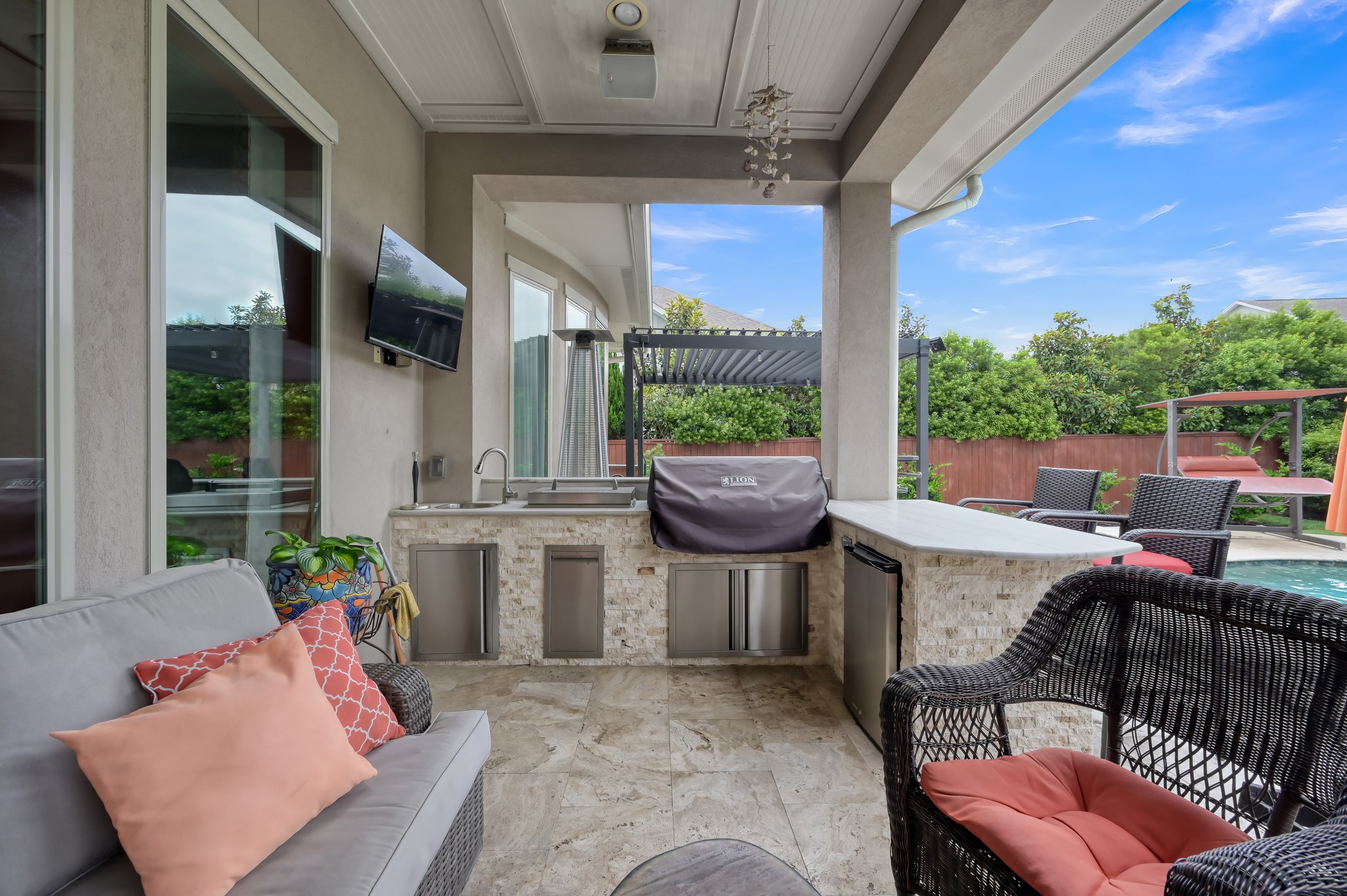 26 Woodborough Way Spring, TX 77389 - Photo 40 of 43 This outdoor living space features a cozy seating area, a built-in barbecue grill, a mini-fridge, and space for a wall-mounted TV. It overlooks a pool, under a shaded pergola, with lush greenery providing privacy and a serene atmosphere.