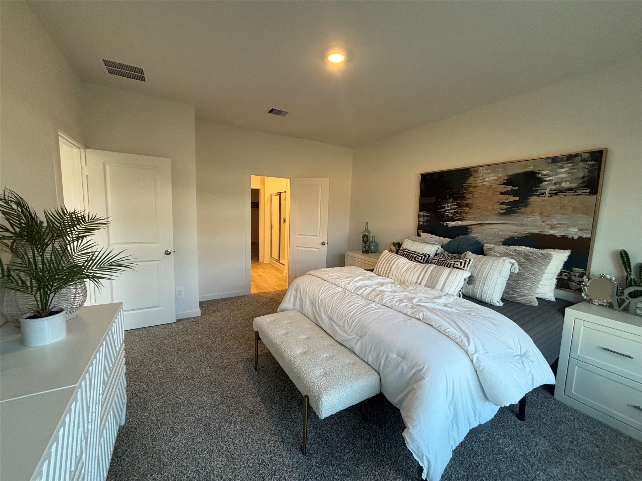 4315 Windsor Forest Fulshear, TX 77494 - Photo 14 of 50 Spacious and modern Primary bedroom on main floor with plush carpeting, featuring a cozy bed, stylish decor, and ample natural lighting. There's an ensuite bathroom and a chic dresser with a plant accent, creating a comfortable and inviting space.