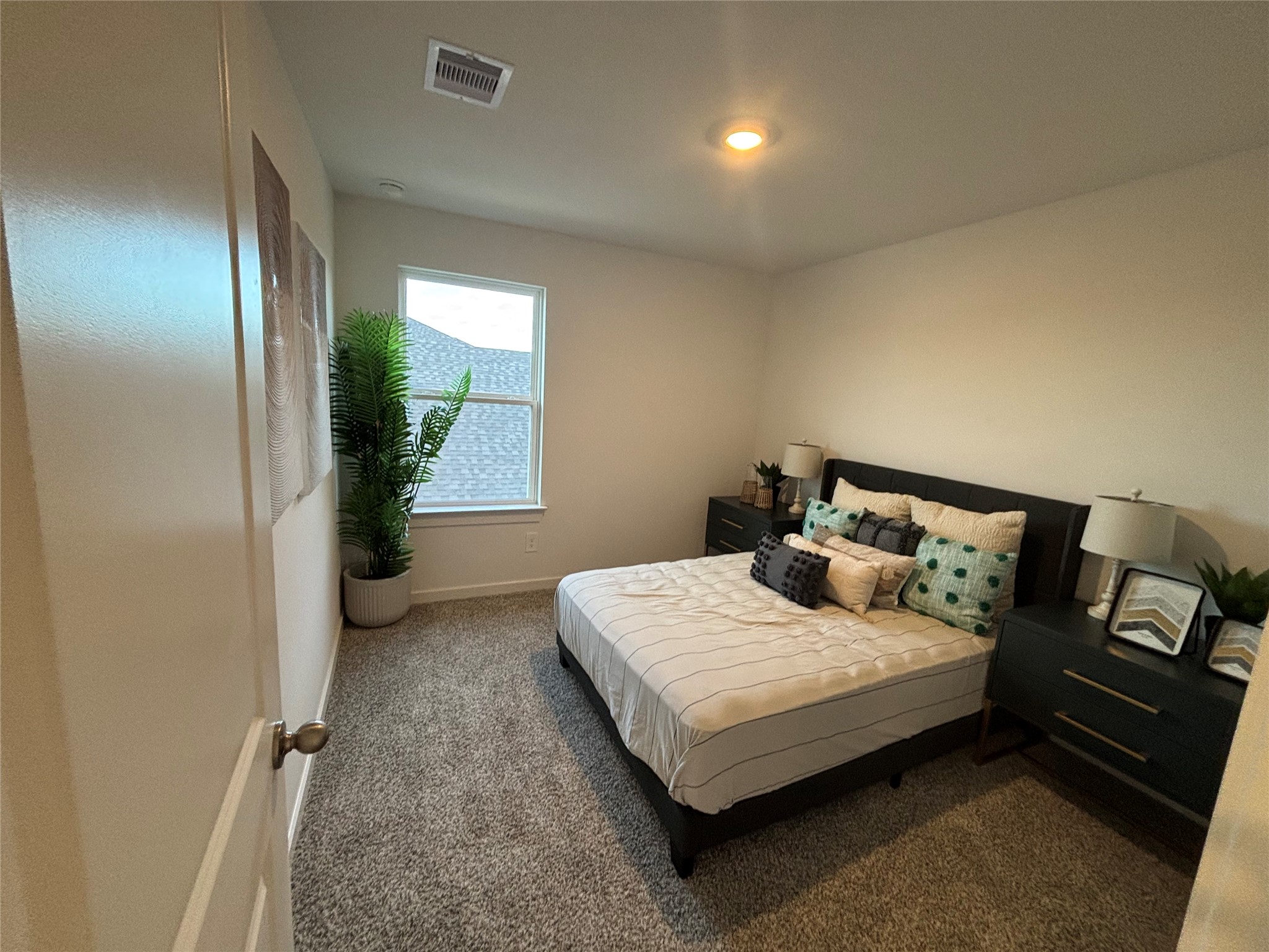 4315 Windsor Forest Fulshear, TX 77494 - Photo 29 of 50 This bedroom features soft carpeting, a modern bed with decorative pillows, and matching nightstands with lamps. The room is brightened by a large window and a potted plant, creating a welcoming atmosphere.