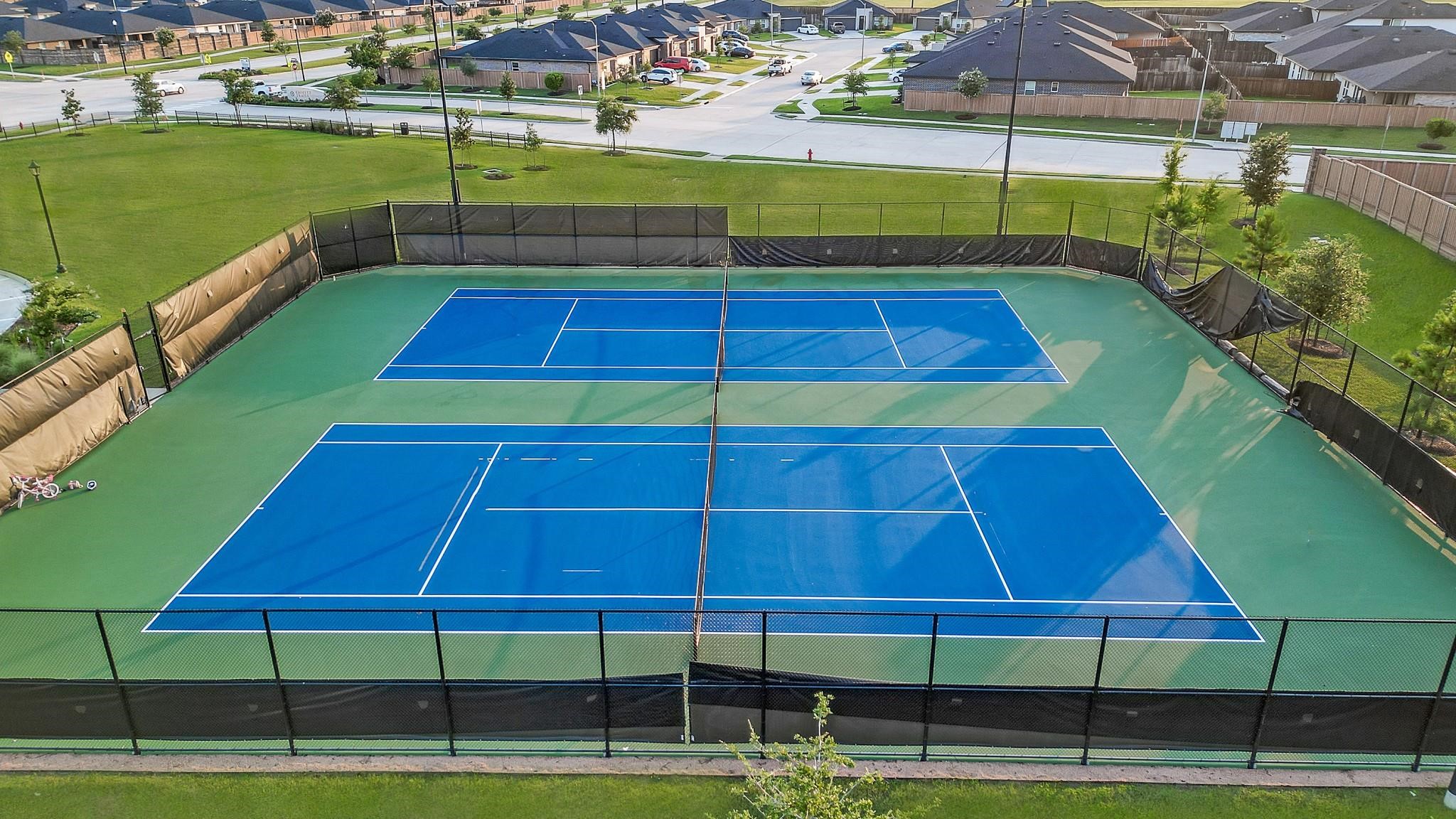 4315 Windsor Forest Fulshear, TX 77494 - Photo 42 of 50 This photo features a well-maintained, double tennis court with blue surfaces and green borders, surrounded by a black fence. It's located in a residential area with nearby homes, offering a great recreational option for residents.