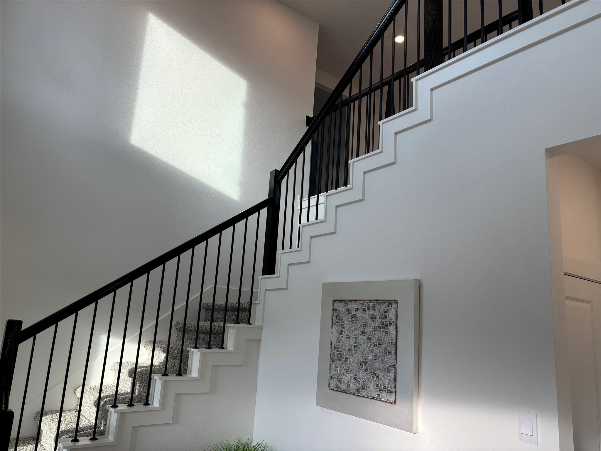 4315 Windsor Forest Fulshear, TX 77494 - Photo 6 of 50 This photo showcases a modern staircase with sleek black railings and carpeted steps, set against a bright, white wall. A piece of abstract art decorates the wall, and natural light enhances the airy feel of the space.