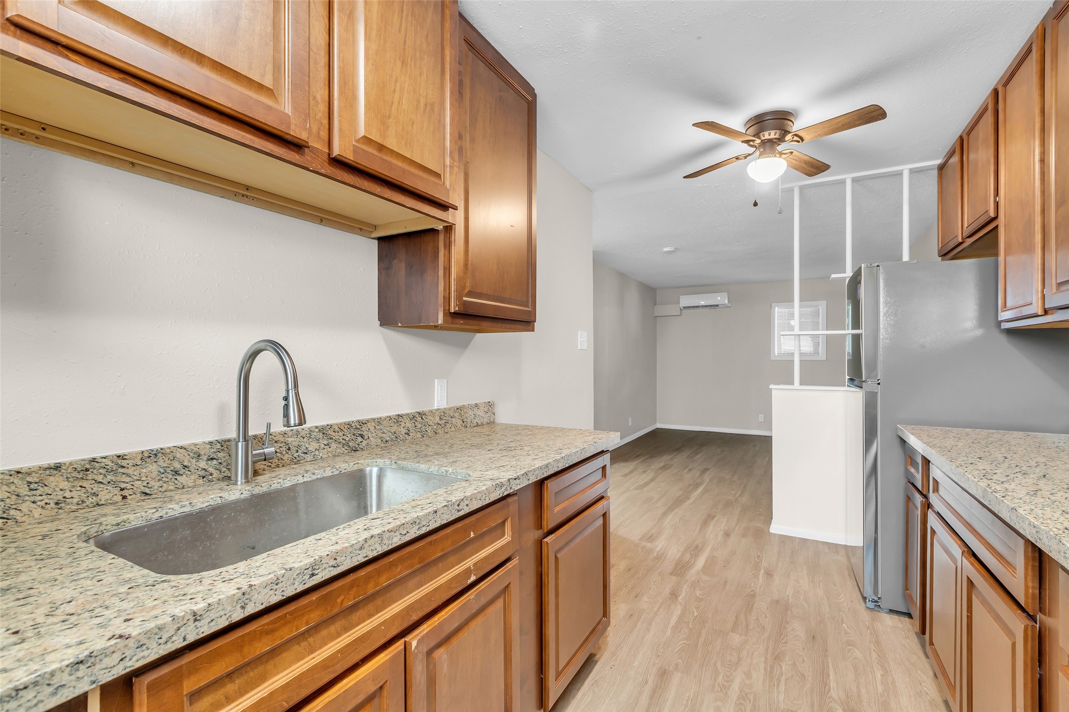 Bright kitchen with granite countertops, wooden cabinets, and stainless steel appliances. Features an open layout with a ceiling fan and wood-style flooring, leading into a spacious living area.