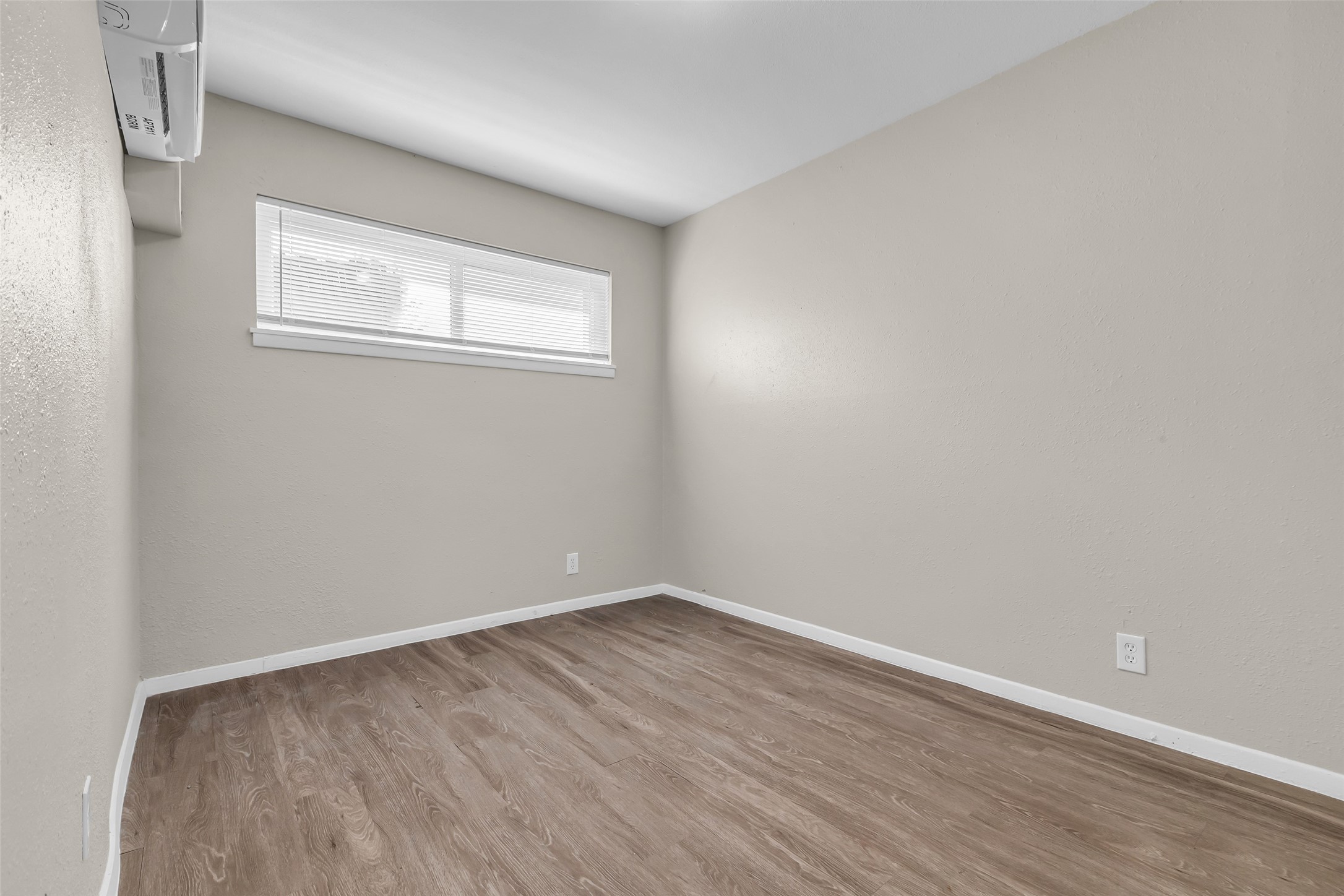 4706 Yellowstone Boulevard, Unit 5 Houston, TX 77021 - Photo 5 of 14 This photo shows a small, unfurnished room with light beige walls and wood-style flooring. It has a narrow window with blinds and a wall-mounted air conditioning unit.