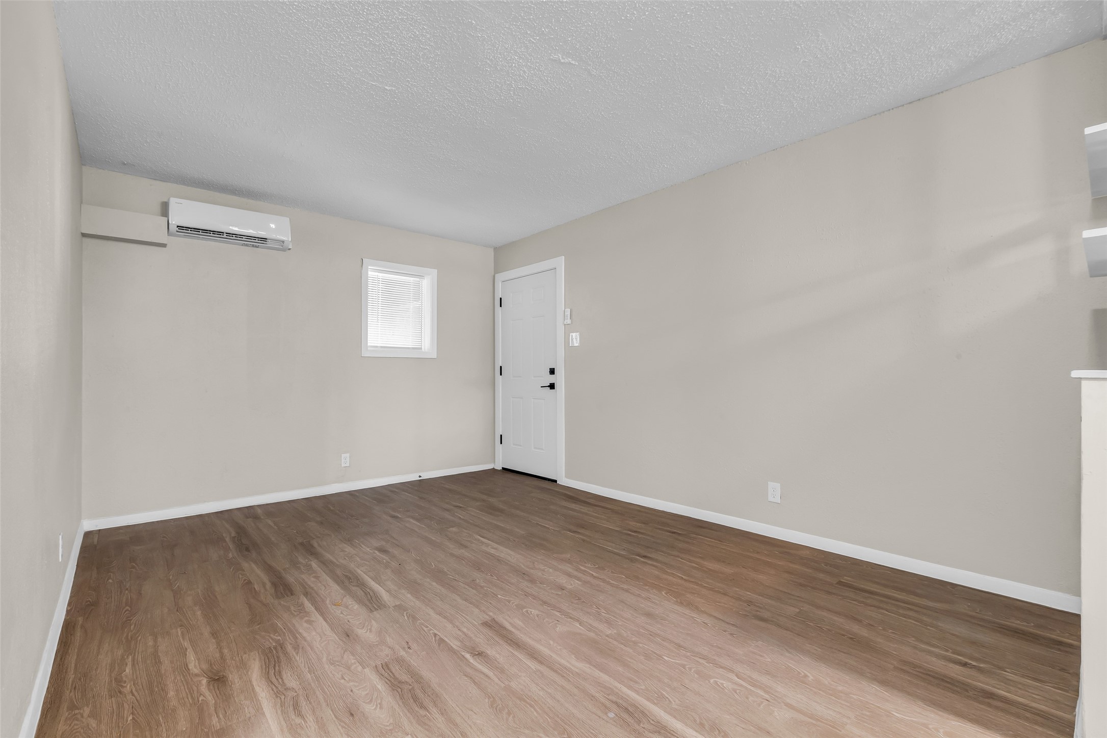 4706 Yellowstone Boulevard, Unit 5 Houston, TX 77021 - Photo 7 of 14 This bright room features hardwood flooring, neutral walls, a small window, and a wall-mounted air conditioning unit. It has a simple, open layout with a white door leading outside.