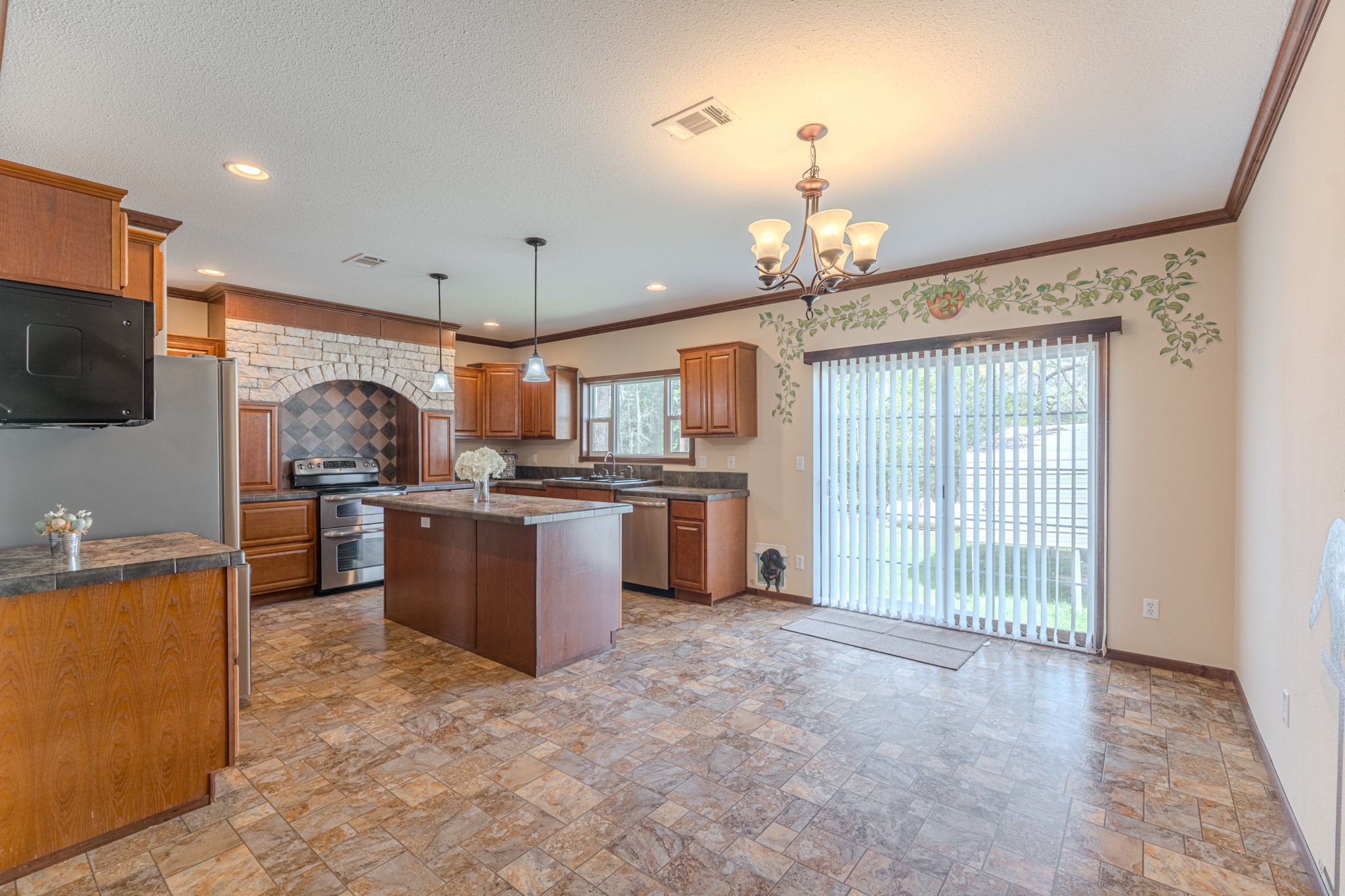 115 Canadian Drive Trinity, TX 75862 - Photo 14 of 41 Kitchen features warm wood cabinetry, a central island, and stainless steel appliances. The room is enhanced by a brick archway above the stove, a chandelier, and sliding glass doors leading to the backyard, providing ample natural light.