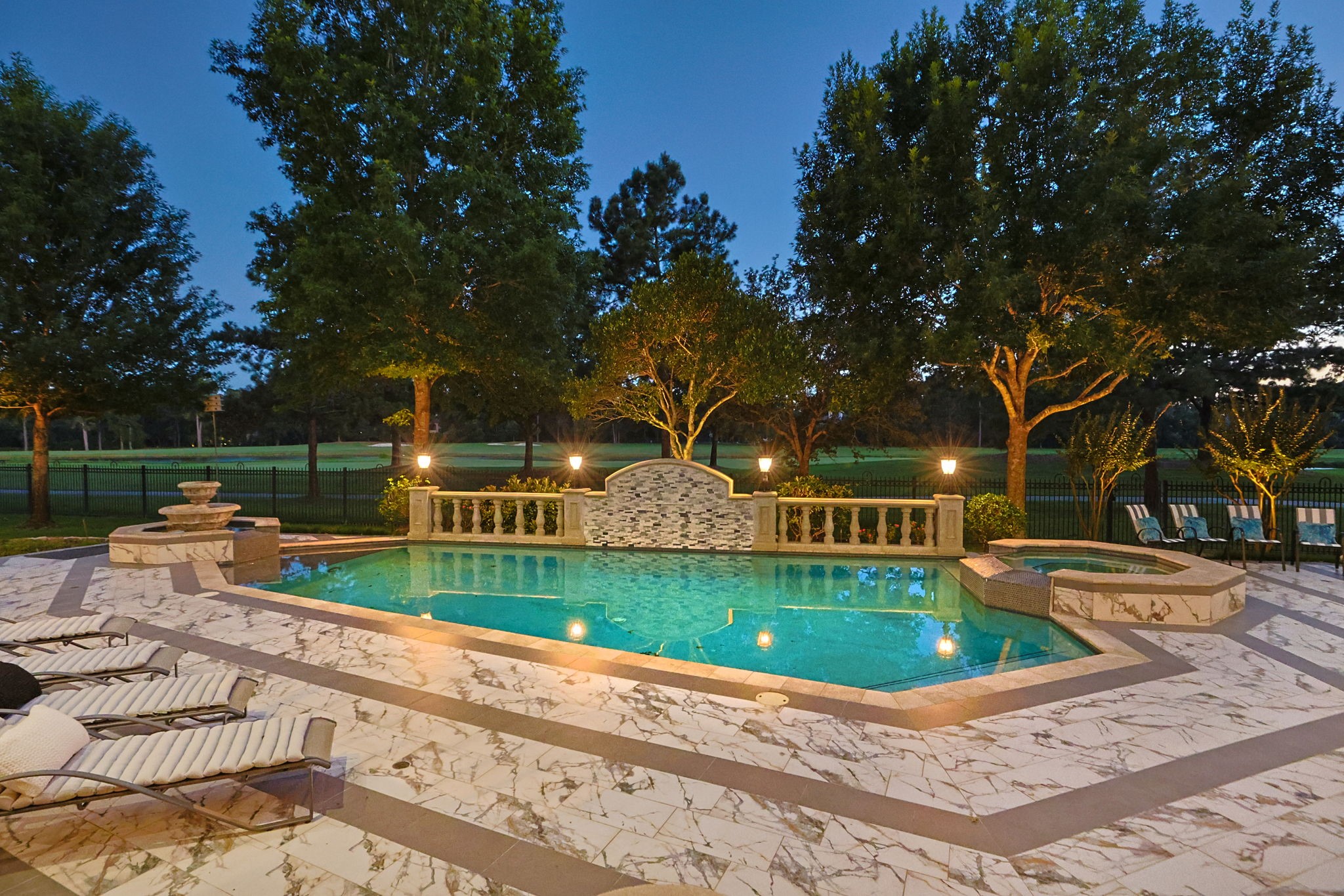 39 Maymont Way Spring, TX 77382 - Photo 42 of 50 This image showcases a luxurious backyard with a beautifully lit swimming pool surrounded by elegant stonework and lush greenery. There are comfortable lounge chairs, a hot tub, and a fountain feature, creating a serene and inviting outdoor space perfect for relaxation and entertaining.
