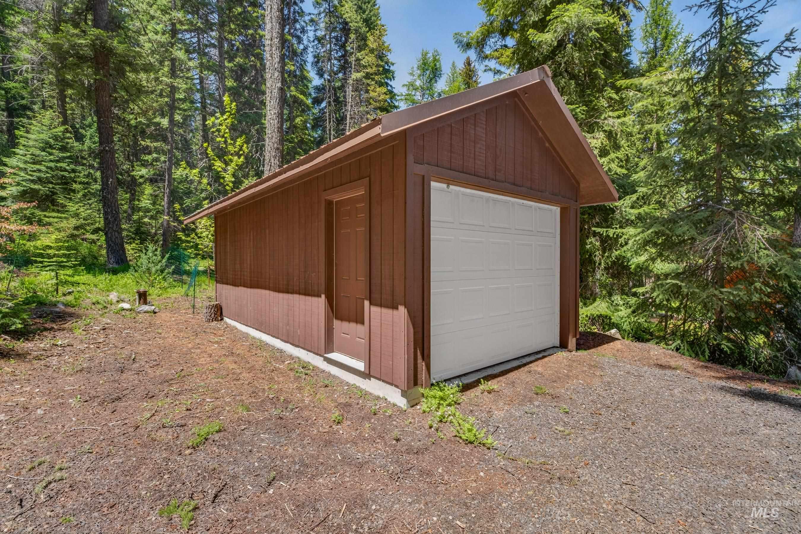 2339 West Mountain Road Donnelly, ID 83615 - Photo 33 of 37 Single-car garage with a brown wood-paneled exterior, white overhead door, and a separate side entry door, set on a concrete foundation