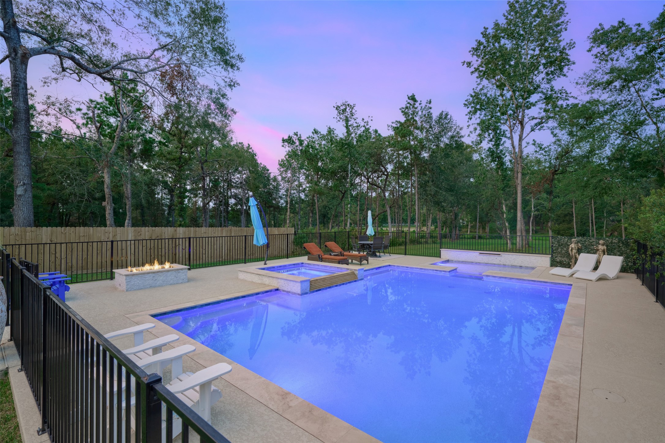 2218 Honea Egypt Road Magnolia, TX 77354 - Photo 22 of 50 This photo showcases a serene backyard with a modern swimming pool and integrated spa, an inviting fire pit area, and lounging spaces. The property is bordered by a privacy fence and lush trees, creating a tranquil outdoor retreat.