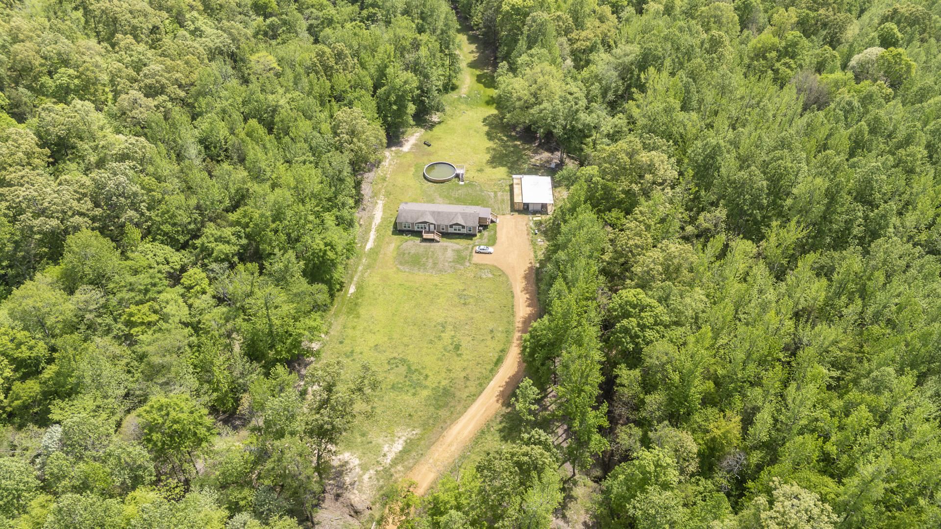 Expansive wooded acreage featuring a single-story residence, a detached auxiliary building, a circular above-ground pool, a clear access road, and a substantial cleared area surrounding the structures
