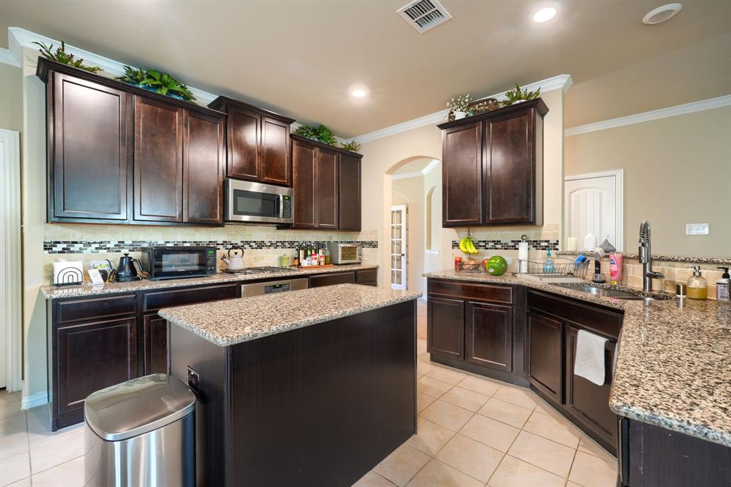 12417 Hitch Rack Way McKinney, TX 75071 - Photo 7 of 17 Kitchen featuring light stone counters, backsplash, ornamental molding, dark brown cabinets, and light tile floors