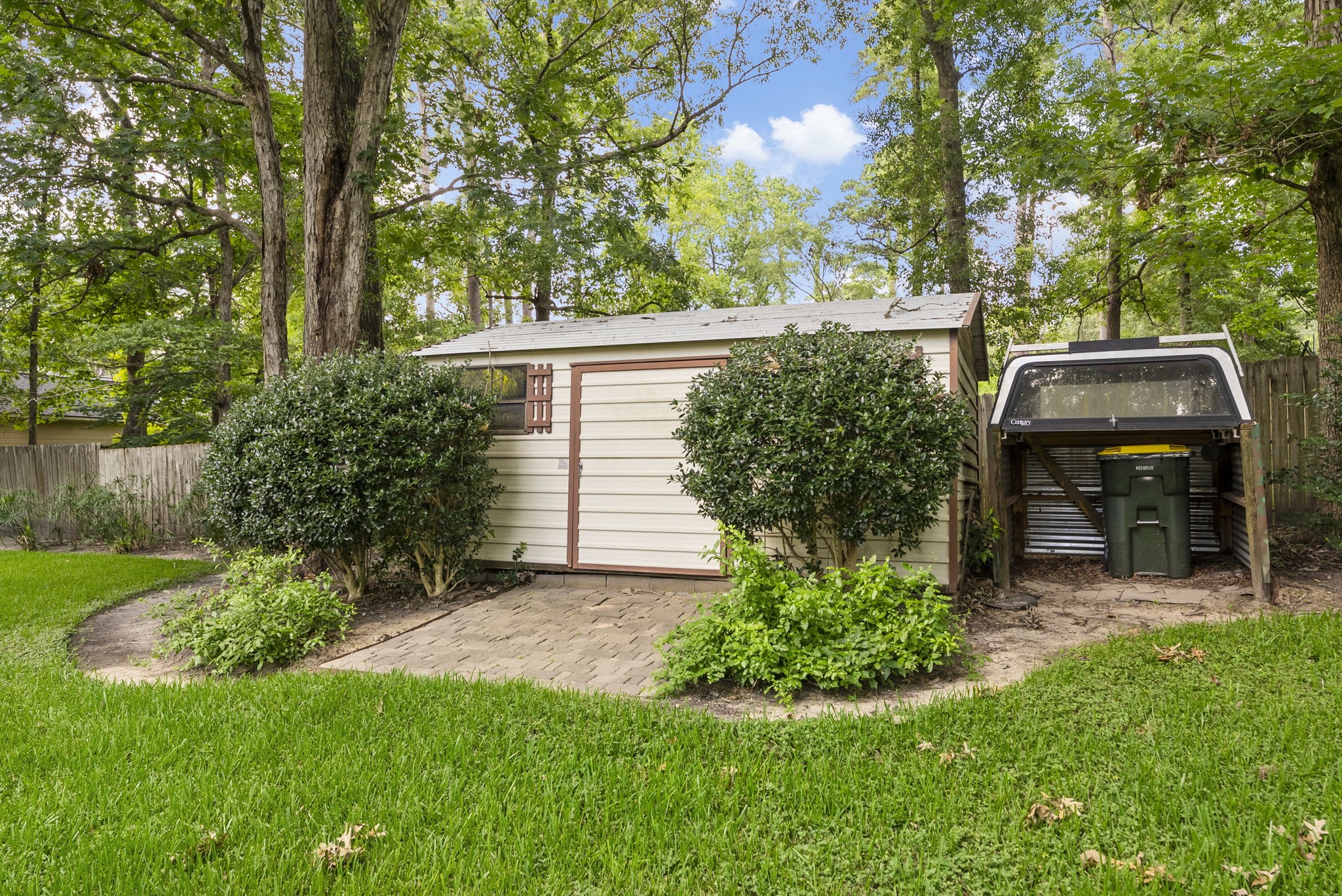 202 Sunset Boulevard Conroe, TX 77303 - Photo 40 of 50 This photo shows a backyard with a small storage shed, surrounded by lush greenery and mature trees. The area appears well-maintained, with a paved path leading to the shed, and a separate covered space for trash bins.