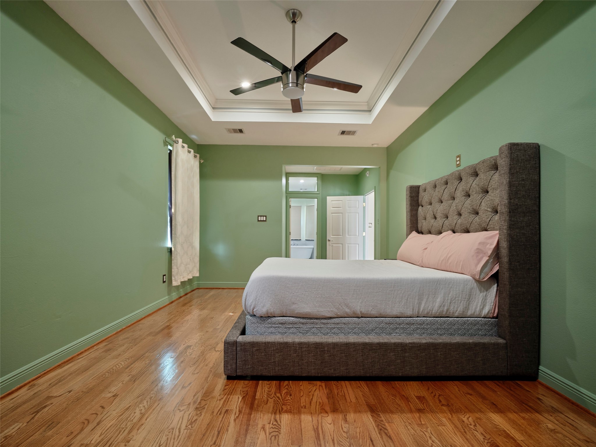 4219 Redbud Street Houston, TX 77051 - Photo 18 of 25 Discover the inviting comfort of this master suite, featuring elegant wood flooring, soothing green walls, a stylish tray ceiling with a modern fan, and a plush, tufted bed for ultimate relaxation.
