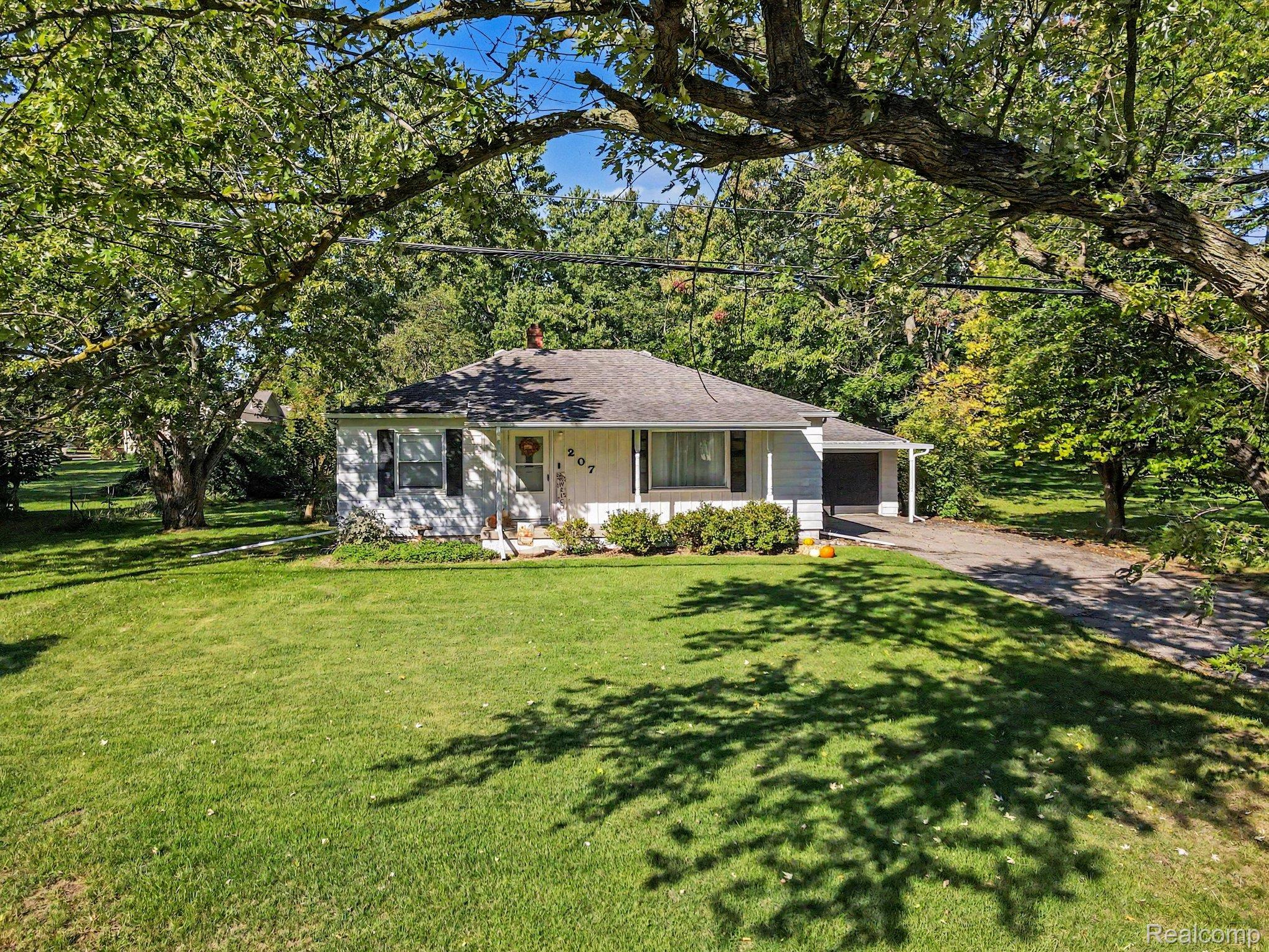 207 Elms Road Flushing, MI 48433 - Photo 27 of 30 Single-story residence with a white exterior, covered entry, front lawn, mature trees, and an attached carport