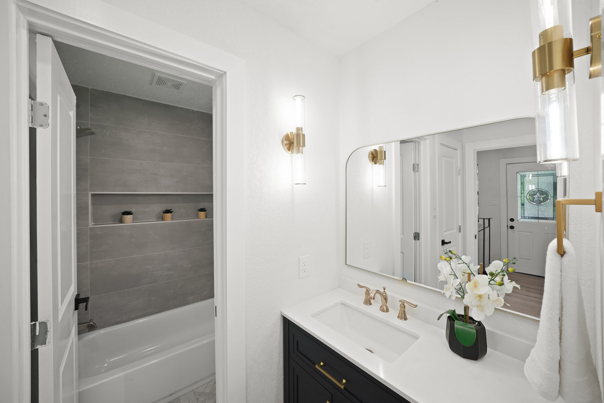 4813 Hummingbird Street Houston, TX 77035 - Photo 31 of 50 The Third Bath (Second Floor of the South Wing) features a Vanity with Quartz Counter & Cabinets/Drawers below, White Undermount Sink, Champagne Bronze Style Faucet, Dual Wall Mounted Brass Style Edison Bulb Sconces, Brass Style Framed Mirror, & a Brass Style Towel Ring. Also offered is a Shelved Linen Closet and a One Piece Swiss Madison Commode. Straight Ahead, Notice the Full Tub/Shower with Light Metallic Grey Format Tile Surround, Built in Shampoo Shelf, Rain Shower Head, and LED Flush Mount.