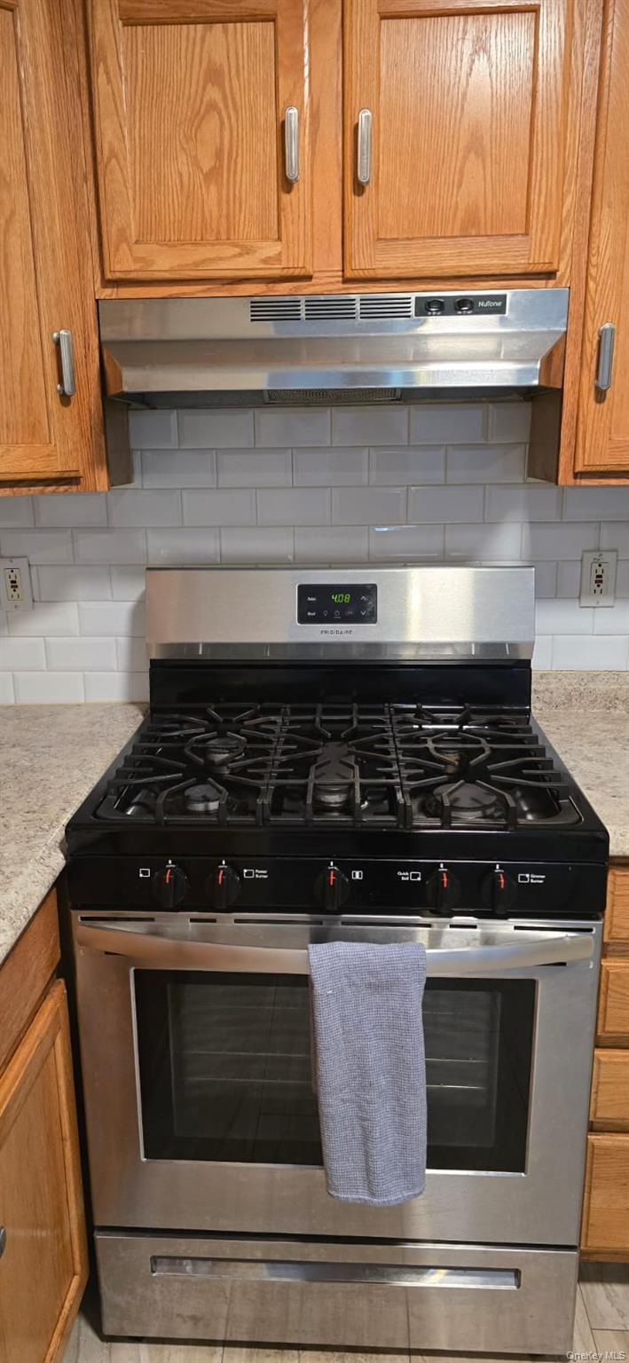 235 MacDougal Street Brooklyn, NY 11233 - Photo 16 of 21 Kitchen view of brown cabinetry, stainless steel range with gas cooktop, under cabinet range hood, tasteful backsplash, and light stone counters