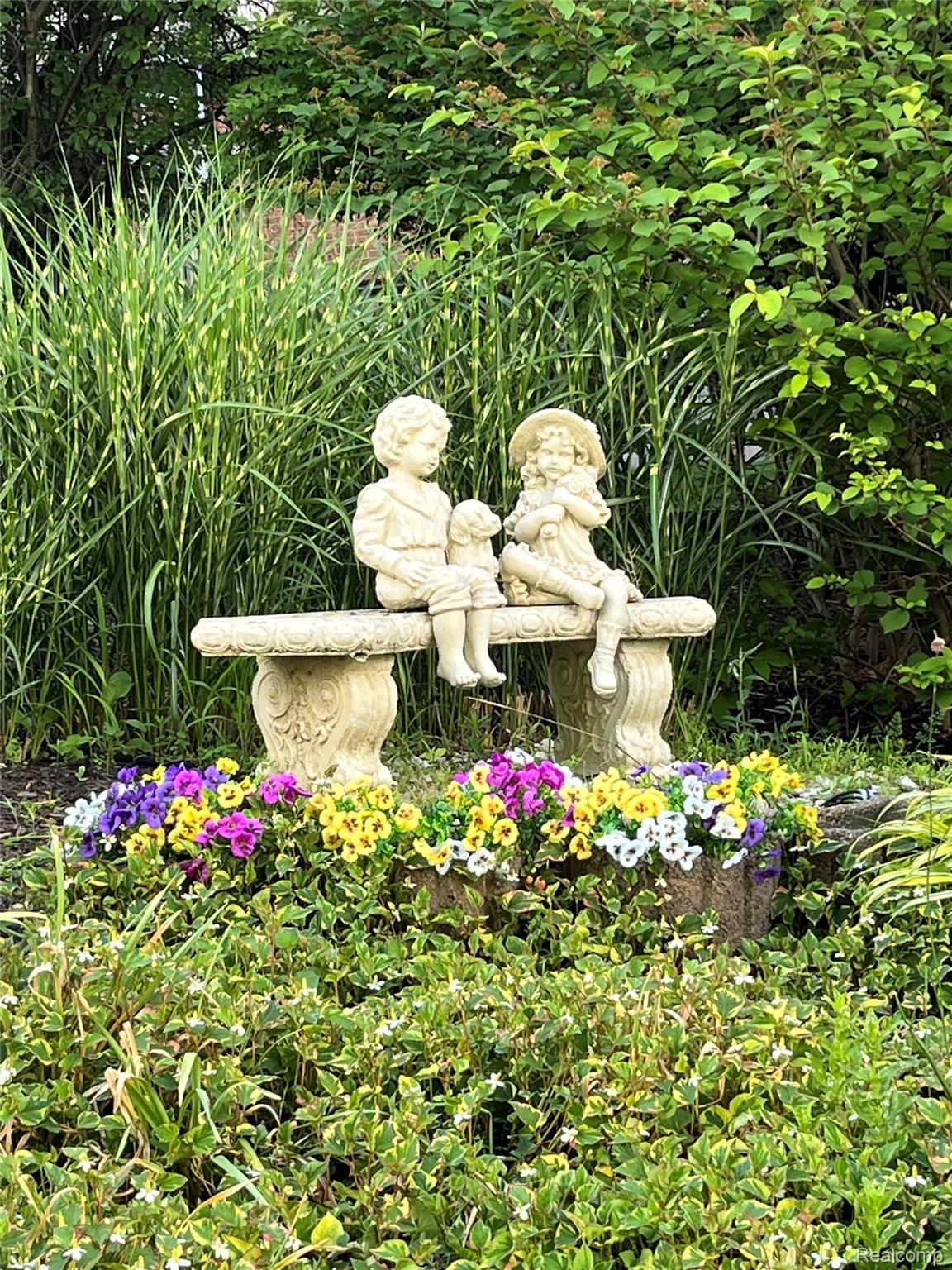 1721 Archers Pointe Rochester, MI 48306 - Photo 6 of 58 Decorative garden bench with statuary, surrounded by lush green foliage, variegated grasses, and vibrant annual flowers