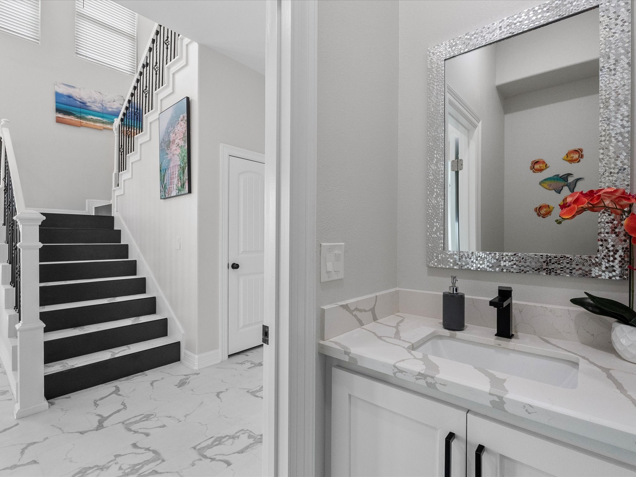 1018 Seashell Drive Surfside Beach, TX 77541 - Photo 32 of 49 This photo shows a modern, bright interior with a stylish staircase featuring black steps and white railings. The adjacent bathroom has a sleek design with a marble countertop, decorative mirror, and artistic wall accents. The floor is finished with elegant white and gray tiles, enhancing the home's contemporary look.