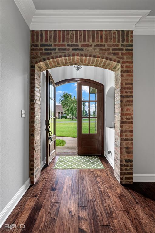 2220 Forest Hills Boulevard Haughton, LA 71037 - Photo 8 of 40 Arched brick doorway featuring transom and sidelight windows on a paneled wood entry door, complemented by wood-finish flooring, white baseboards, and crown molding