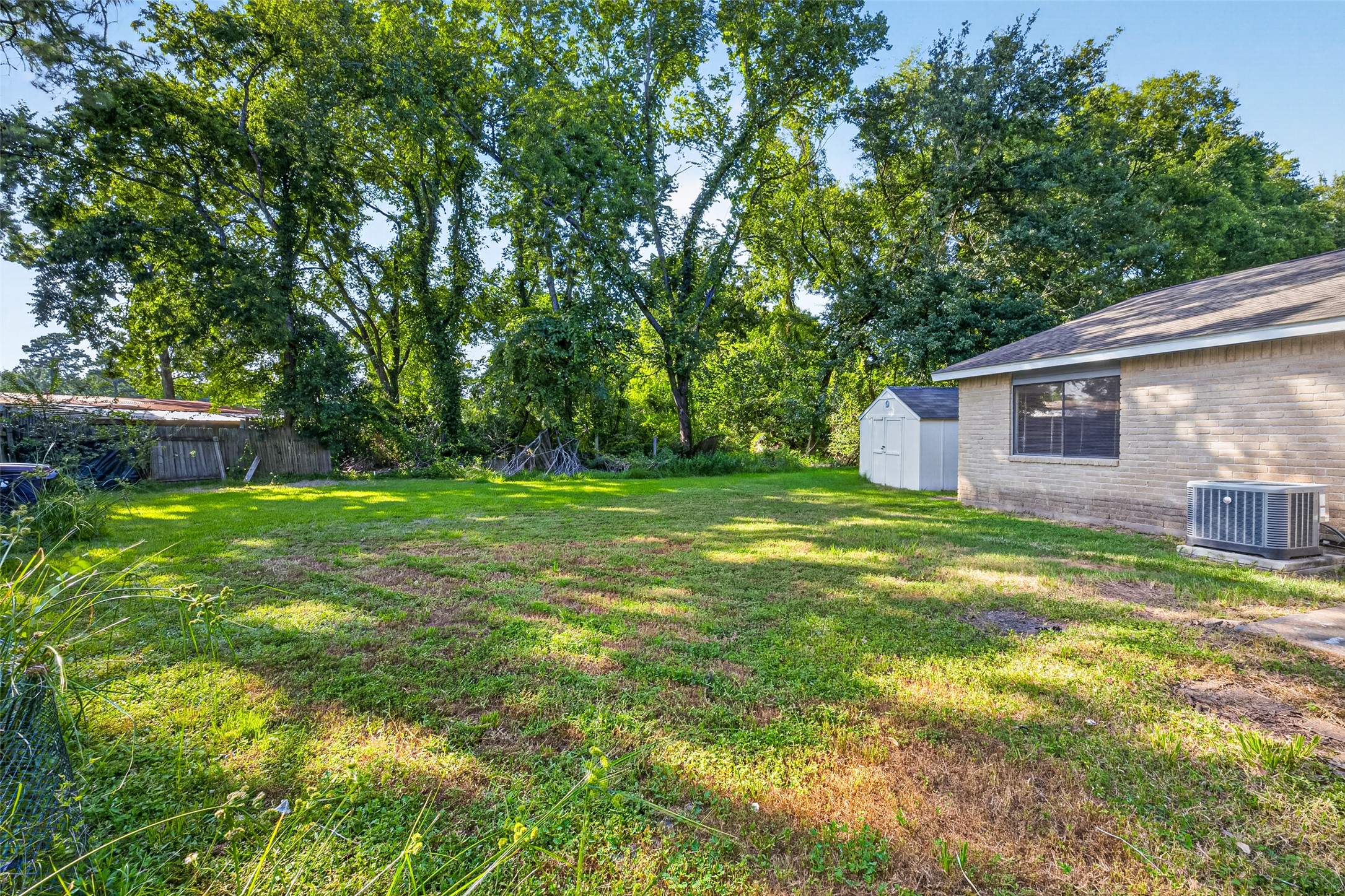 22926 Millgate Drive Spring, TX 77373 - Photo 28 of 34 Spacious backyard with lush grass and mature trees, offering privacy and shade. Includes a small storage shed and is adjacent to a brick house with a large window and air conditioning unit. Ideal for outdoor activities and relaxation.