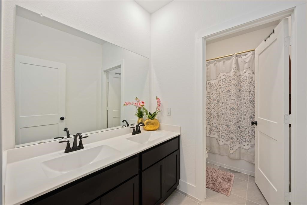 10014 Nectar Path Iowa Colony, TX 77583 - Photo 20 of 26 This bathroom features a modern double vanity with dark cabinetry and sleek fixtures, a large mirror, and a shower with a patterned curtain. The space is bright and inviting with white walls and tile flooring.