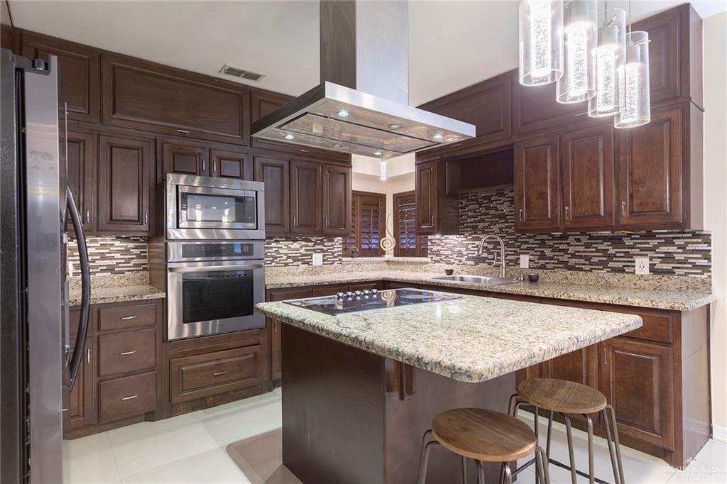 2001 Quail Lane Mission, TX 78572 - Photo 7 of 28 Kitchen featuring a central island with integrated cooktop, granite countertops, dark wood-finish cabinetry, stainless steel appliances, and a mosaic tile backsplash