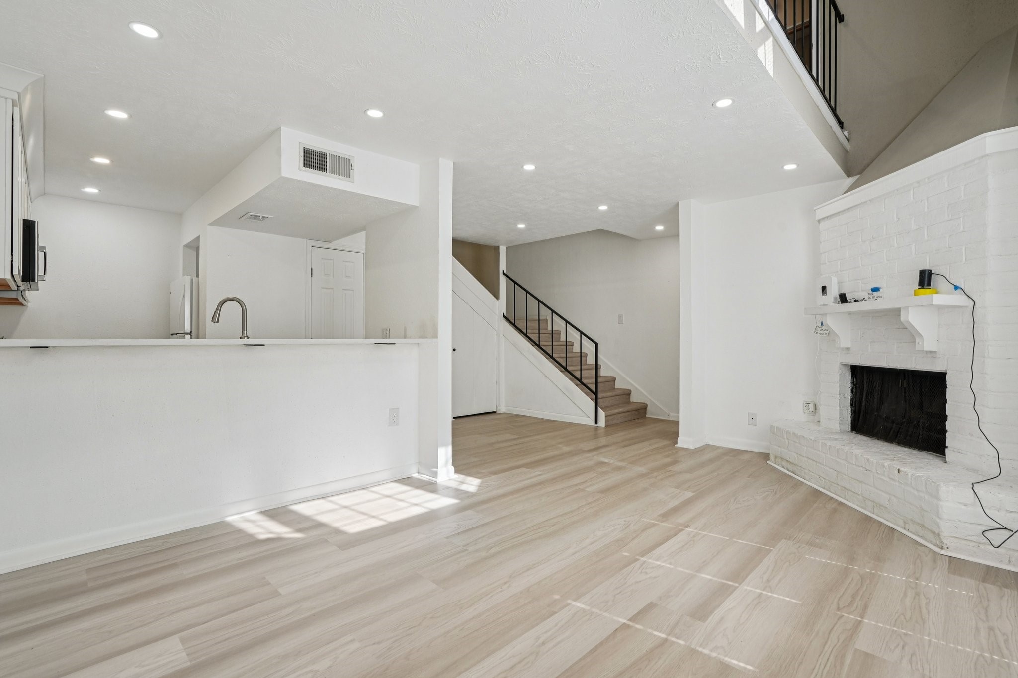 2800 Jeanetta Street, Unit 203 Houston, TX 77063 - Photo 6 of 34 This bright, open-concept living area features modern wood flooring, a sleek white kitchen with a breakfast bar, and a cozy white brick fireplace. The room extends to a staircase leading to the upper level, creating an inviting space.