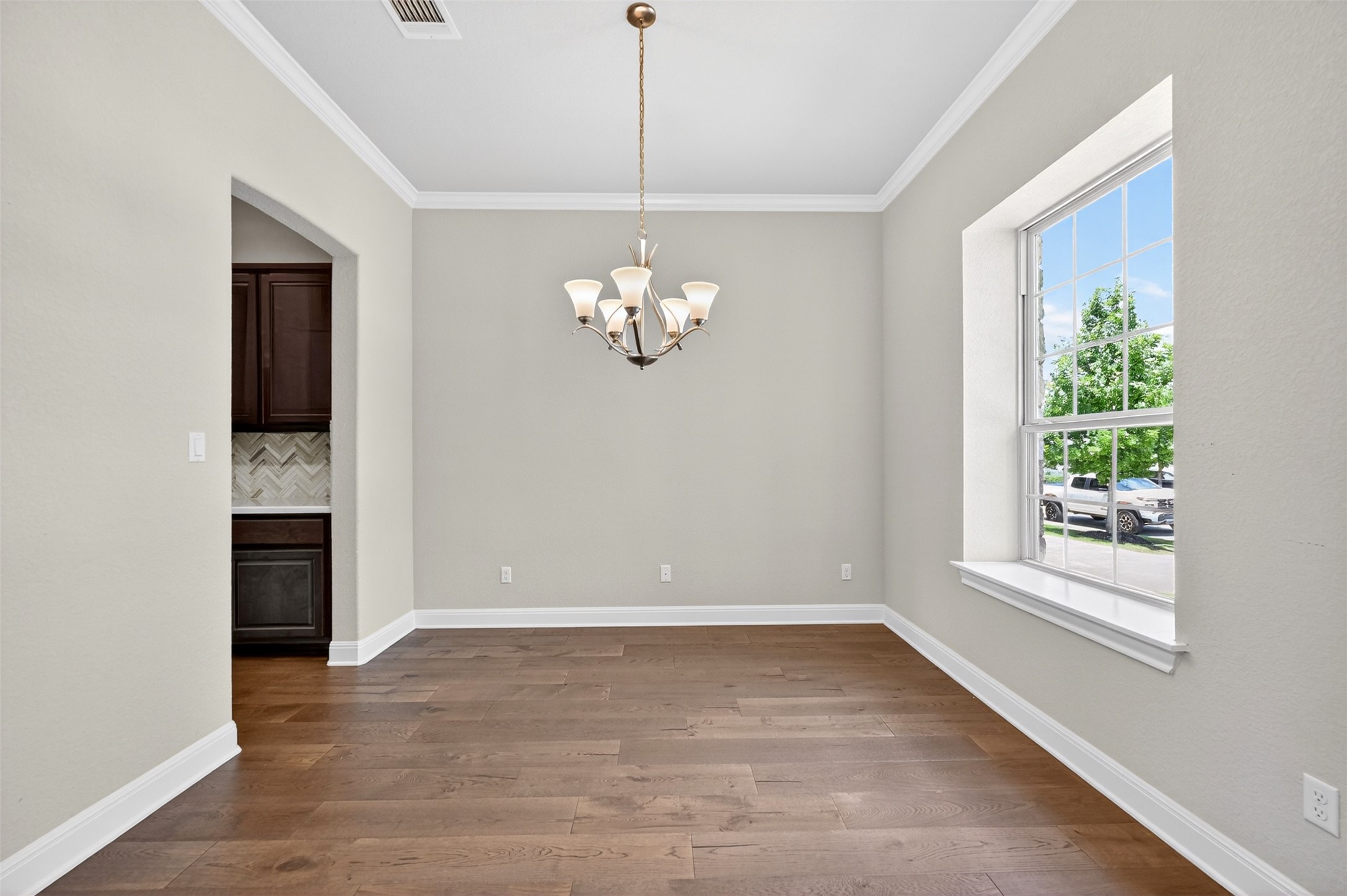 1201 Positano Pass Leander, TX 78641 - Photo 4 of 40 Spacious room with wood-finish flooring, a multi-light chandelier, a large window, crown molding, and an arched entry to a butler's pantry with dark cabinetry and a chevron-patterned tile backsplash