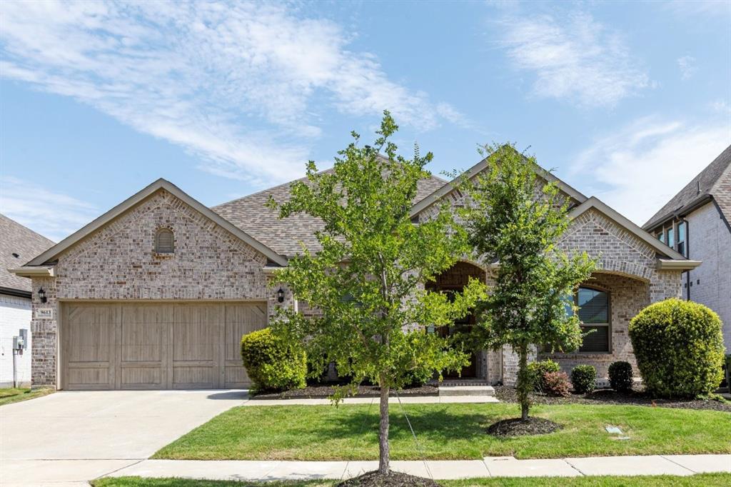 9613 Boulder Pt Road Oak Point, TX 75068 - Photo 3 of 28 Brick facade residence featuring a two-car garage with wood-finish doors, a concrete driveway, and a manicured lawn with mature trees and shrubbery