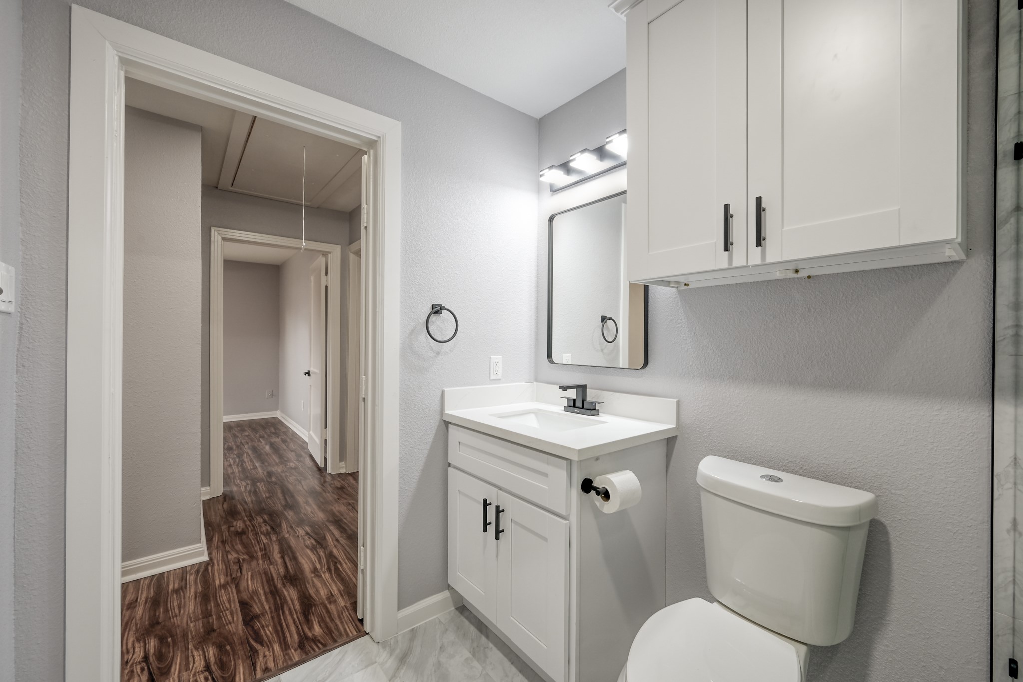 513 Barkley Street Houston, TX 77022 - Photo 17 of 48 This is a modern bathroom with a sleek design, featuring a white vanity and cabinetry, a large mirror, and contemporary fixtures. The light gray walls complement the white accents, and the room has tiled flooring. It leads into a hallway with wood-style flooring.