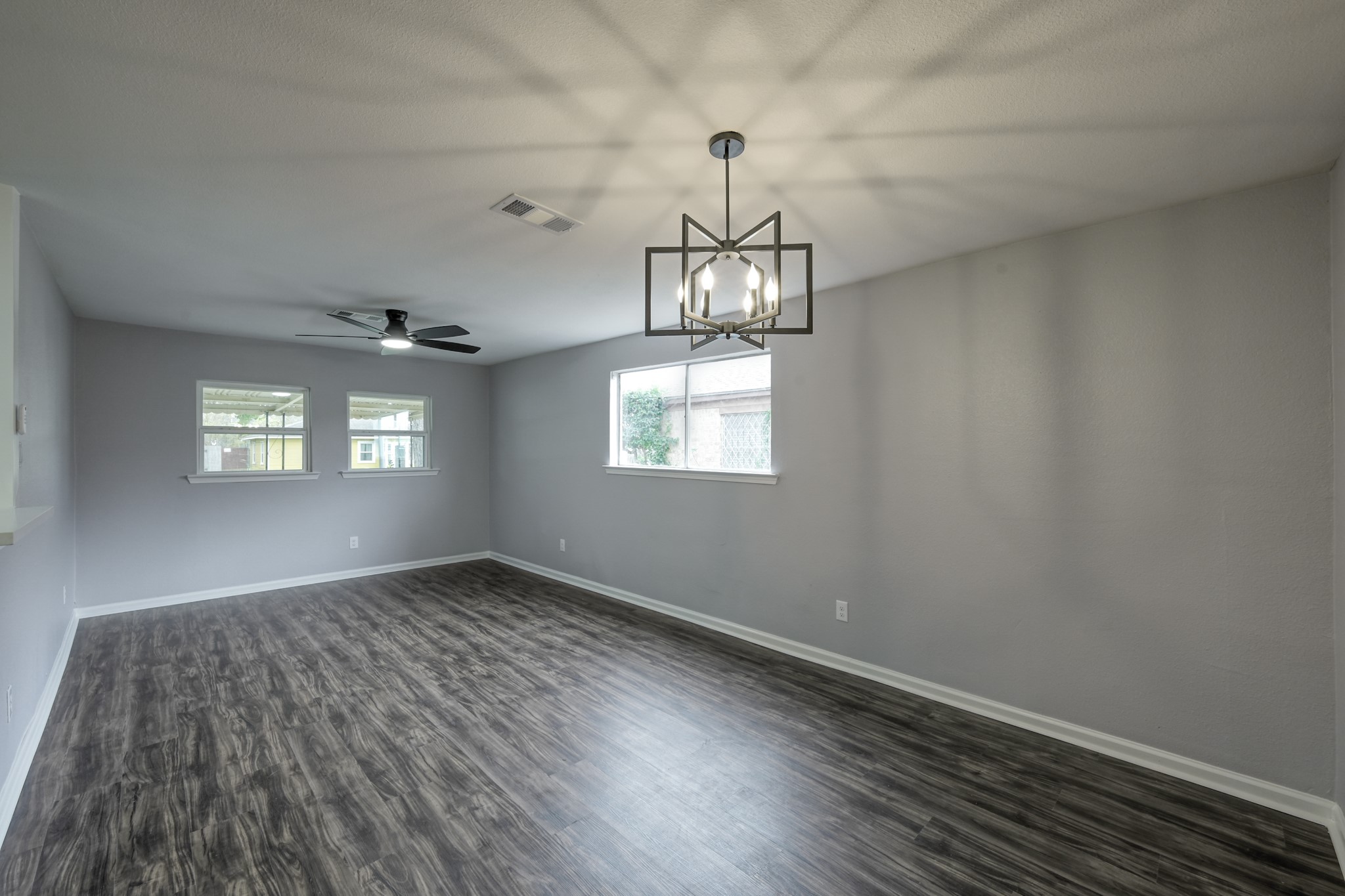 513 Barkley Street Houston, TX 77022 - Photo 37 of 48 This photo showcases a spacious, open room with modern dark wood flooring and light gray walls. It features a stylish, geometric light fixture and a ceiling fan, with ample natural light from two large windows. Ideal for a living or dining area.