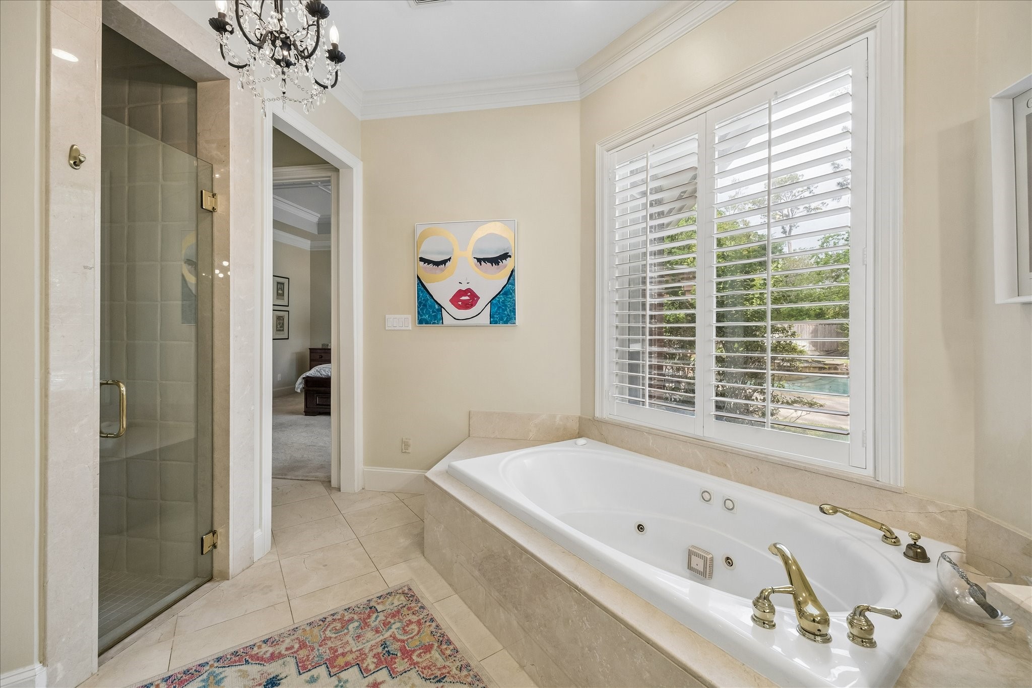 11421 Whippoorwill Road Houston, TX 77024 - Photo 16 of 33 An alternate view of the primary ensuite reveals the frameless glass shower with custom tile work, situated adjacent to the soaking tub. This bright sanctuary features large windows with plantation shutters, neutral stone flooring, and an elegant crystal chandelier, creating a true spa-like atmosphere at home.