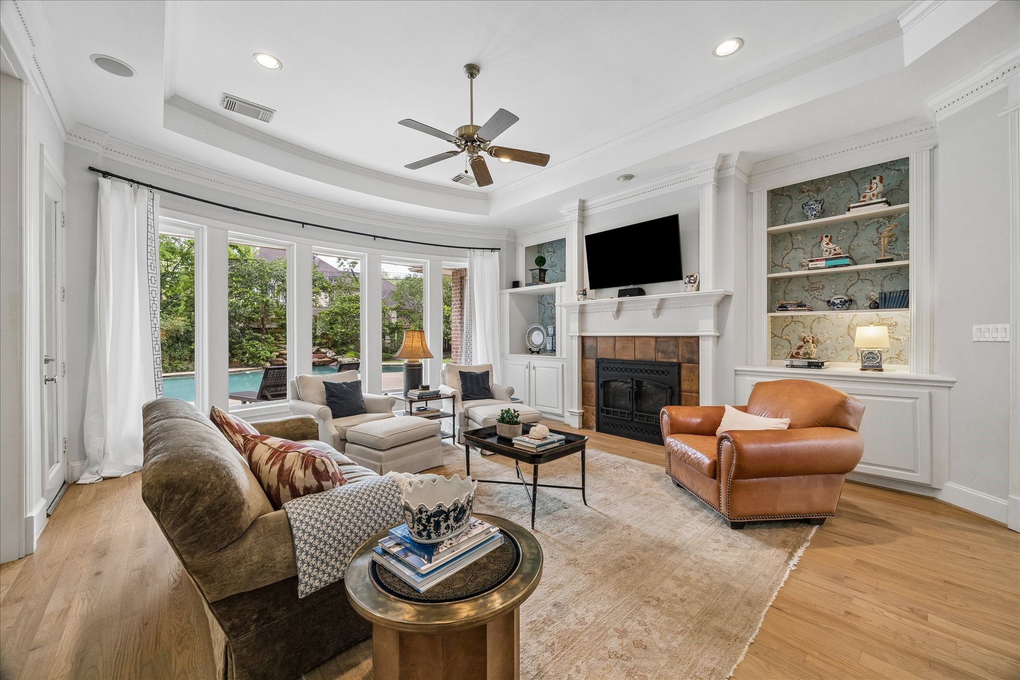 11421 Whippoorwill Road Houston, TX 77024 - Photo 10 of 33 Relax in the light-filled family room, anchored by a curved wall of floor-to-ceiling windows offering serene views of the backyard oasis. This inviting space features a gas-log fireplace, custom built-ins with wallpaper accents, and an elegant double-tray ceiling