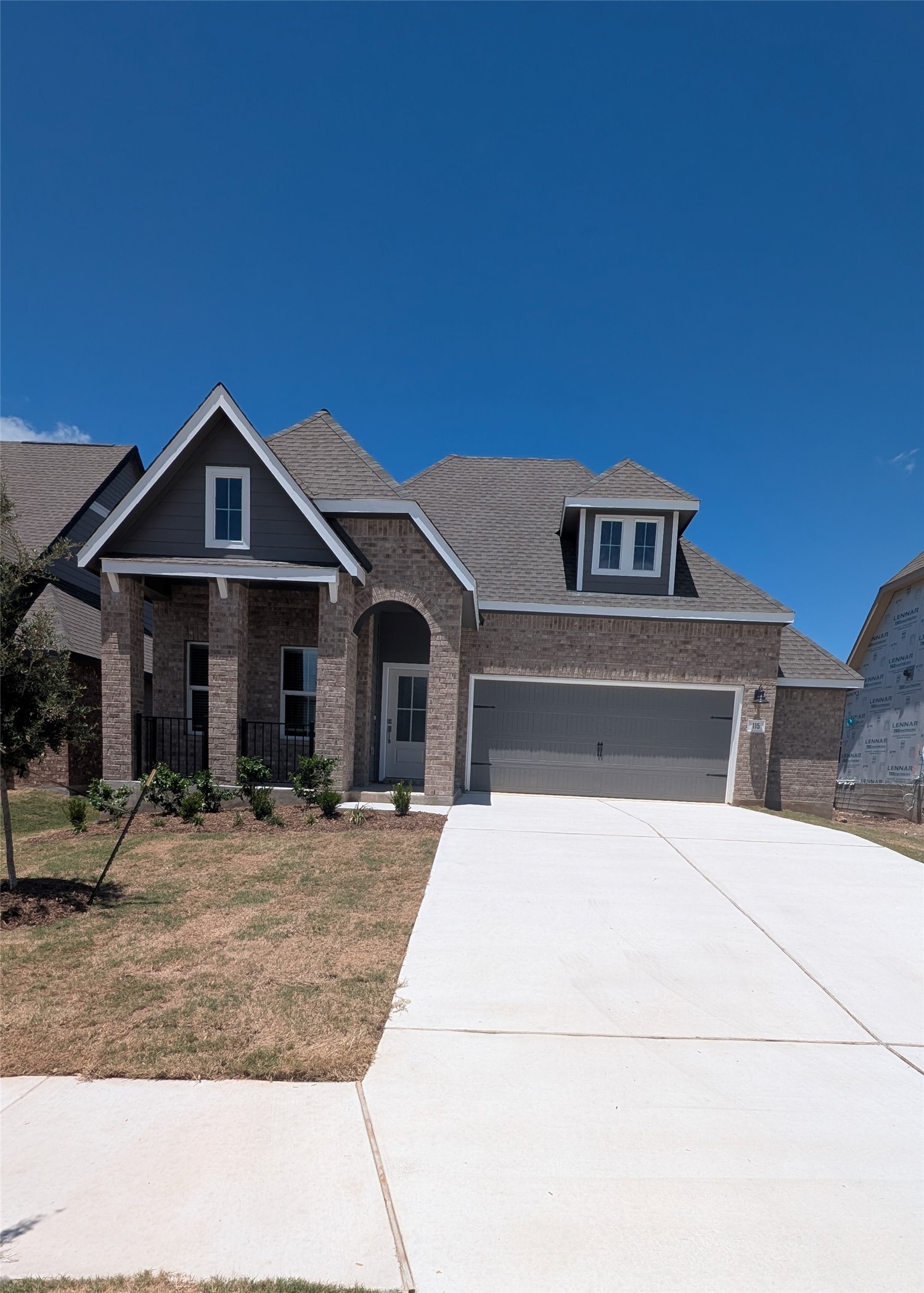 115 Parsons Kyle, TX 78640 - Photo 1 of 2 Brick exterior with gray siding accents, featuring a covered porch entry, multiple dormer windows, an arched entryway, an attached two-car garage, and a concrete driveway