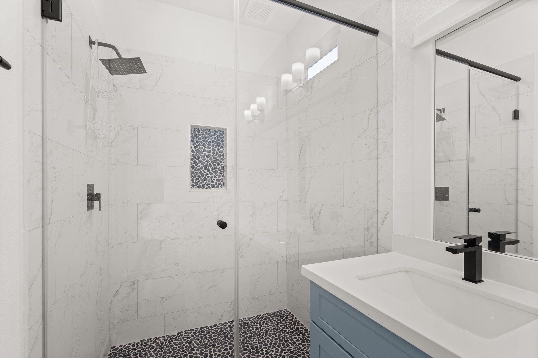 2928 Stevens Street Houston, TX 77026 - Photo 13 of 30 This modern bathroom features a sleek glass-enclosed shower with elegant white marble tiles and a stylish pebble floor. The vanity includes a contemporary sink with a matte black faucet, complemented by a light blue cabinet and a large mirror.