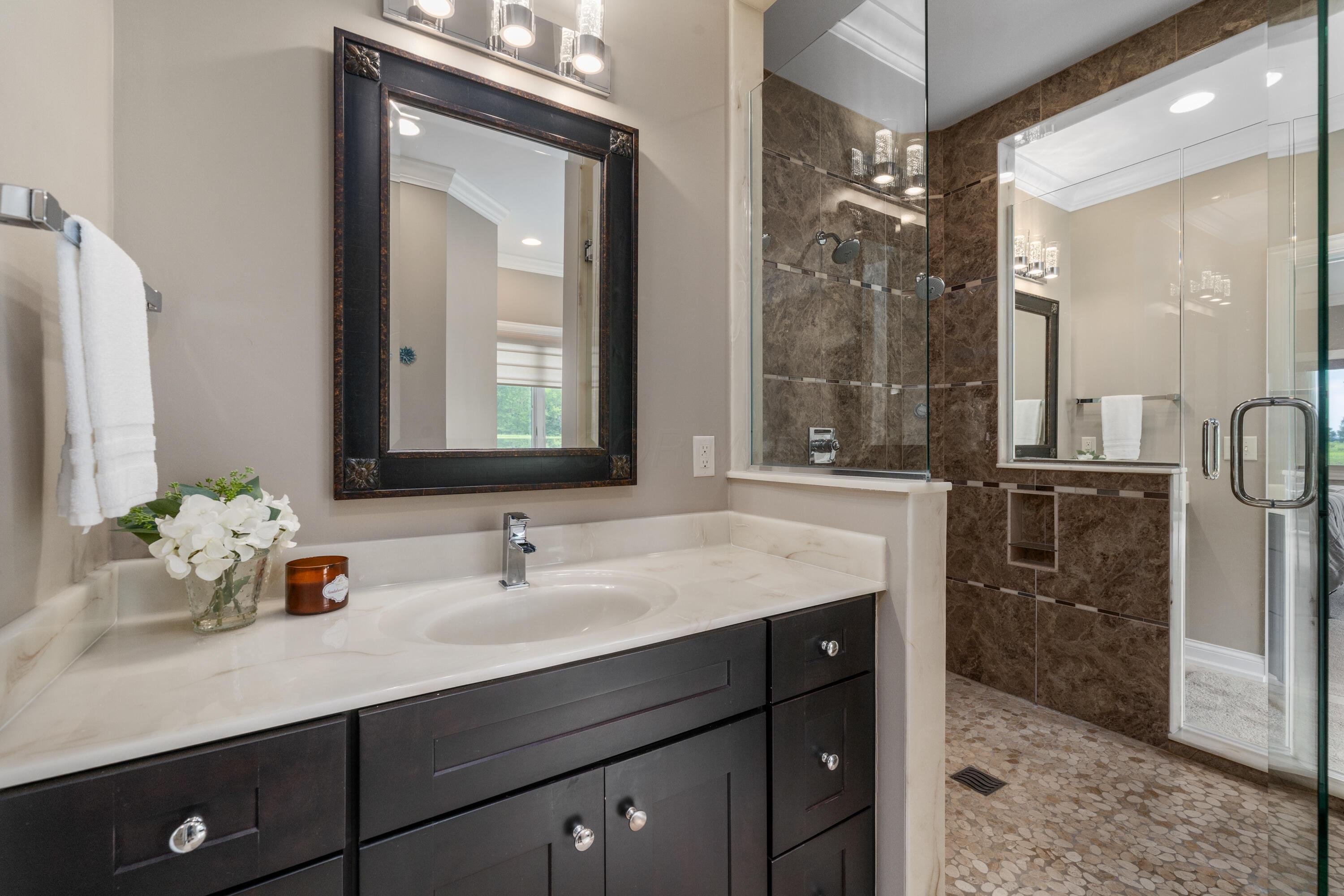 4640 Maynard Road Delaware, OH 43015 - Photo 38 of 91 Bathroom featuring a dark wood-finish vanity with a white countertop and undermount sink, a framed mirror, and a glass-enclosed shower with dark tile walls and pebble flooring
