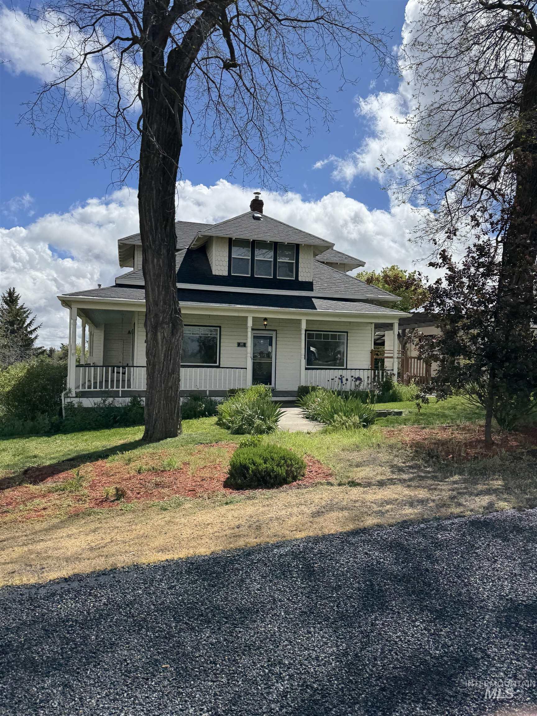 Traditional two-story home featuring a covered front porch with white railing, dormer windows, a prominent front entrance, and a gravel driveway