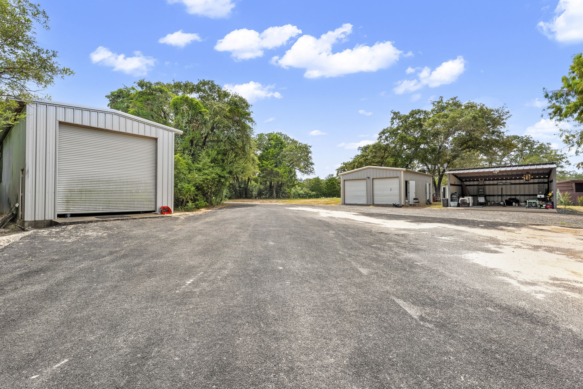 206 22 Hills Road Gause, TX 77857 - Photo 16 of 28 Several detached metal outbuildings flank a wide paved driveway under a blue sky.