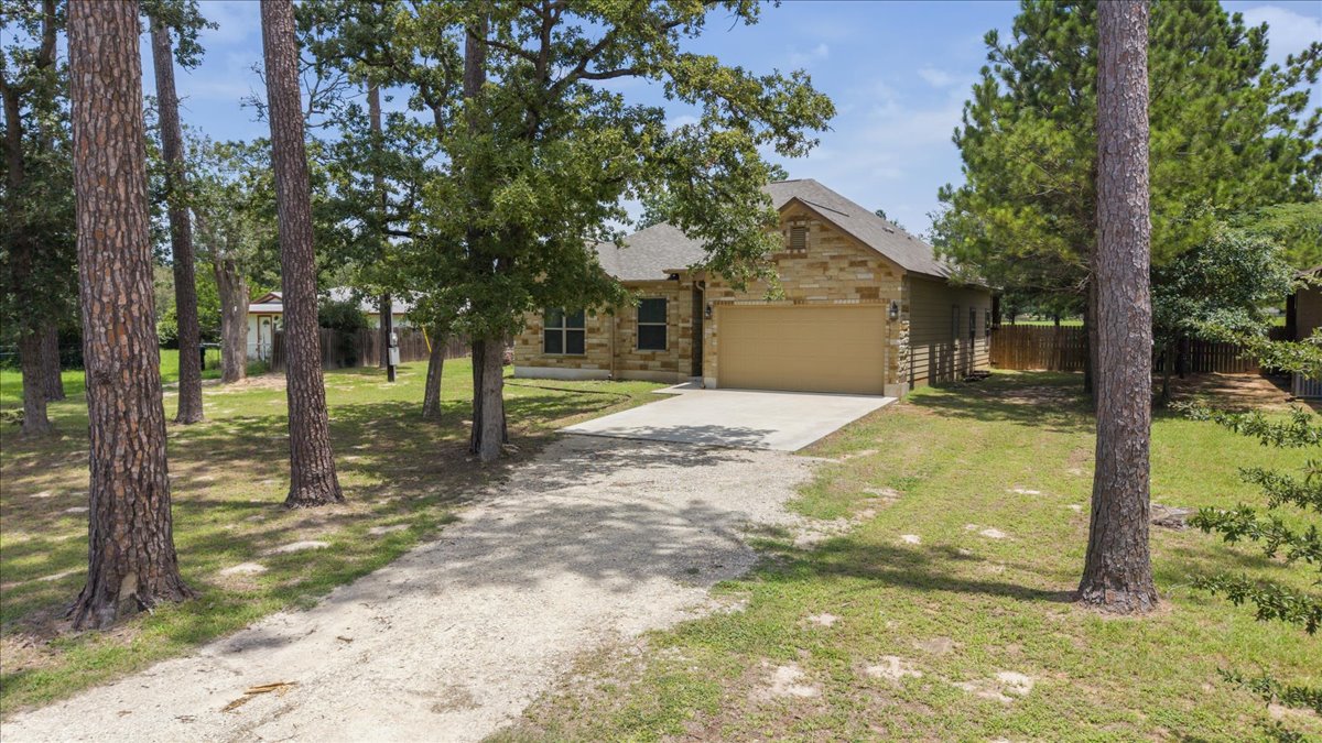 145 Cardinal Loop Paige, TX 78659 - Photo 4 of 40 A welcoming stone facade home nestled among mature trees, featuring a concrete driveway and an attached garage.